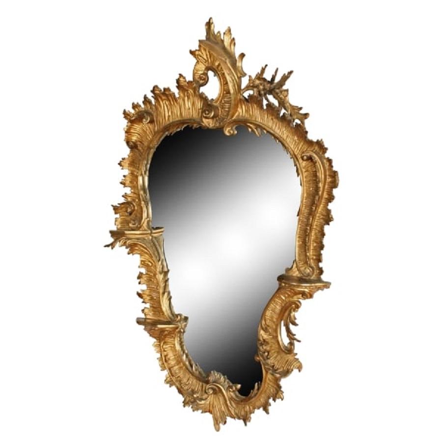 A late 18th to early 19th century carved wood Rococo style gilded mirror.

The shaped carved frame is a true Rococo design with scrolls, flowers and three small platform shelves.

The gilding is original but worn and there are some chips and