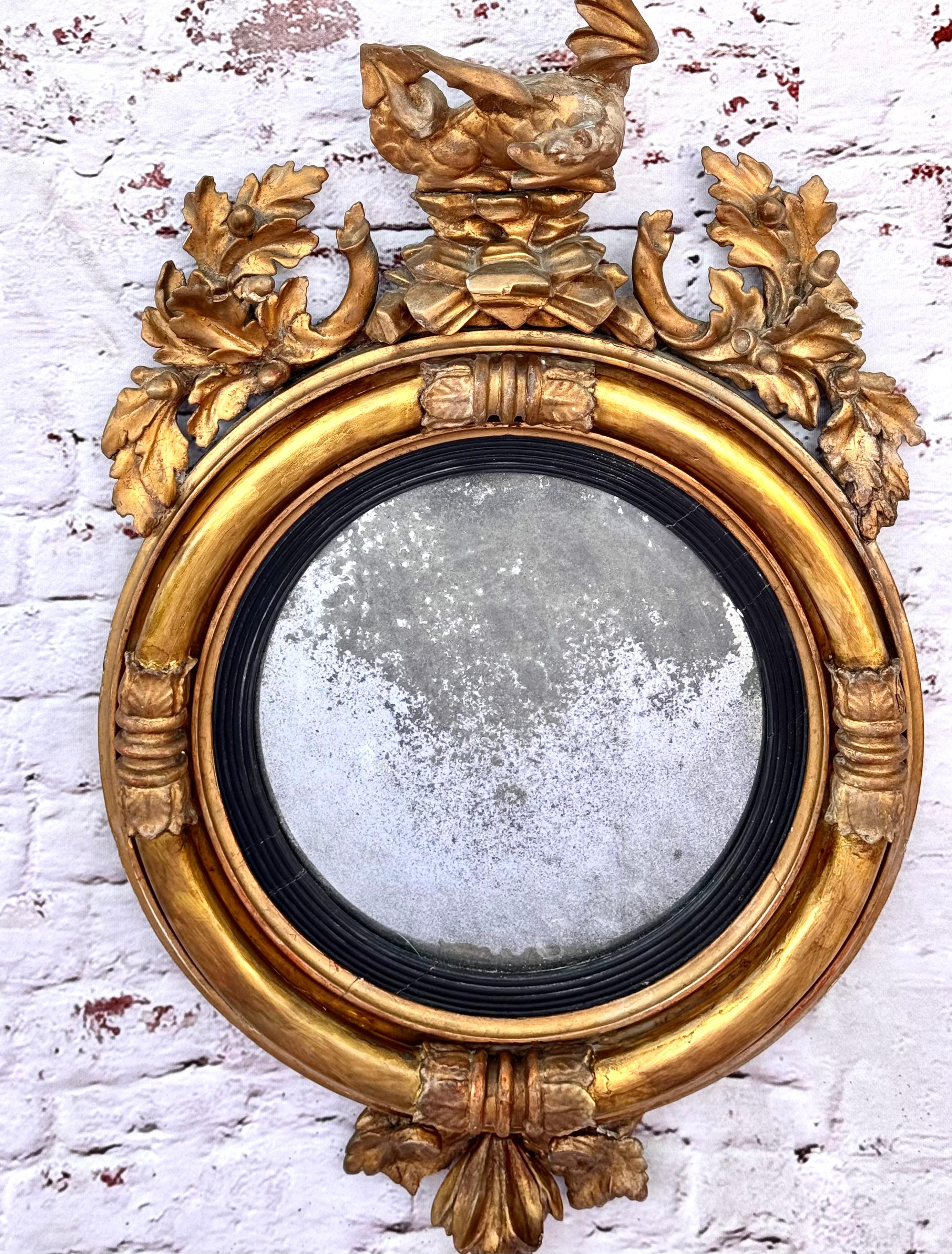 Early 19th century circular regency giltwood convex mirror.  Mirror features a carved dragon perched atop a structure of rocks and a foliate pendant grouping on bottom. The patina on the gilt highlights the carvings of the acanthus leaves along with