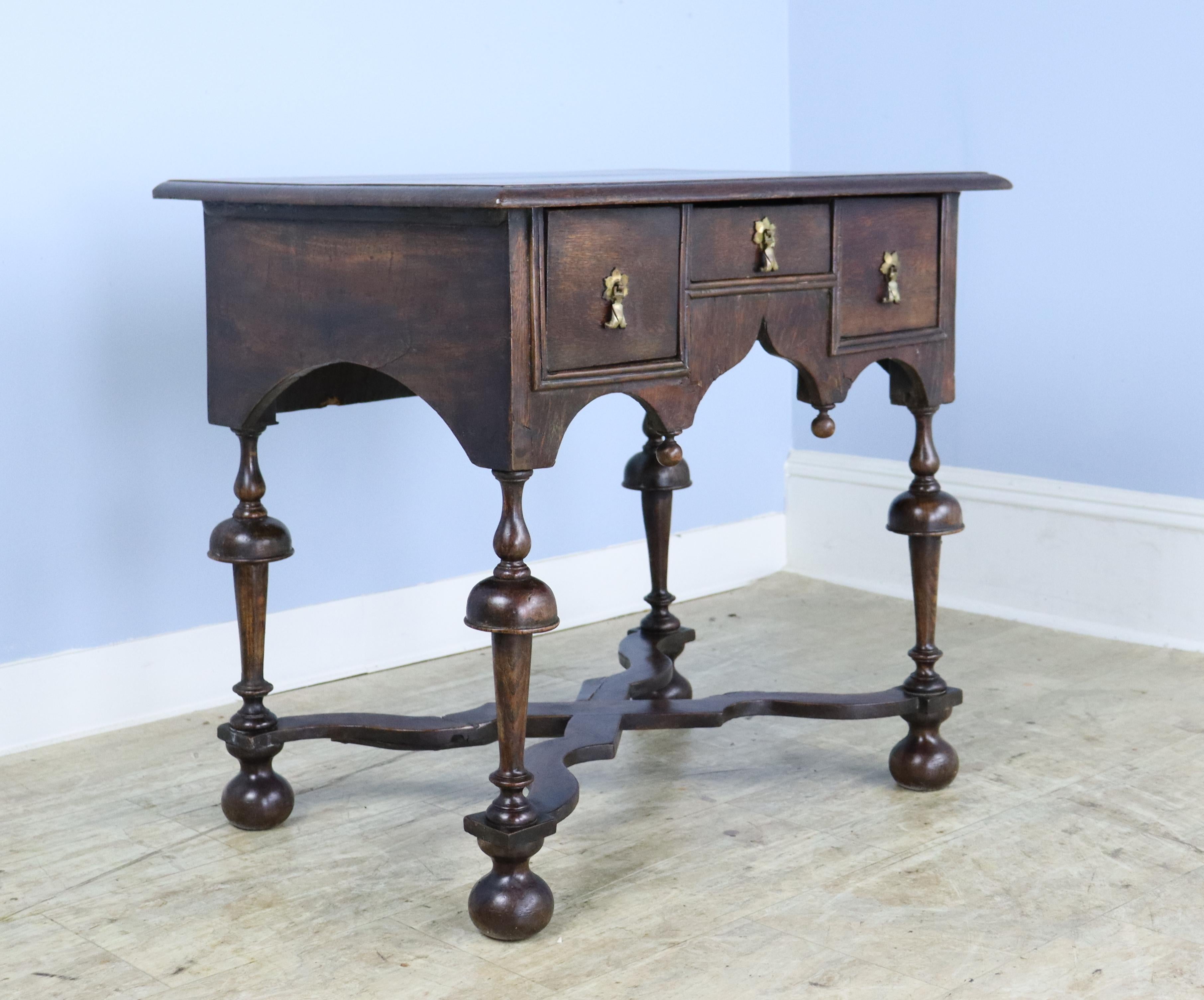 An antique lowboy from England with period bun feet and stylized finials. Three drawers with pretty original drop escutcheons. Note the elaborately carved apron and the unusual carved  criss-cross stretcher base. The oak has a rich and warm color. A