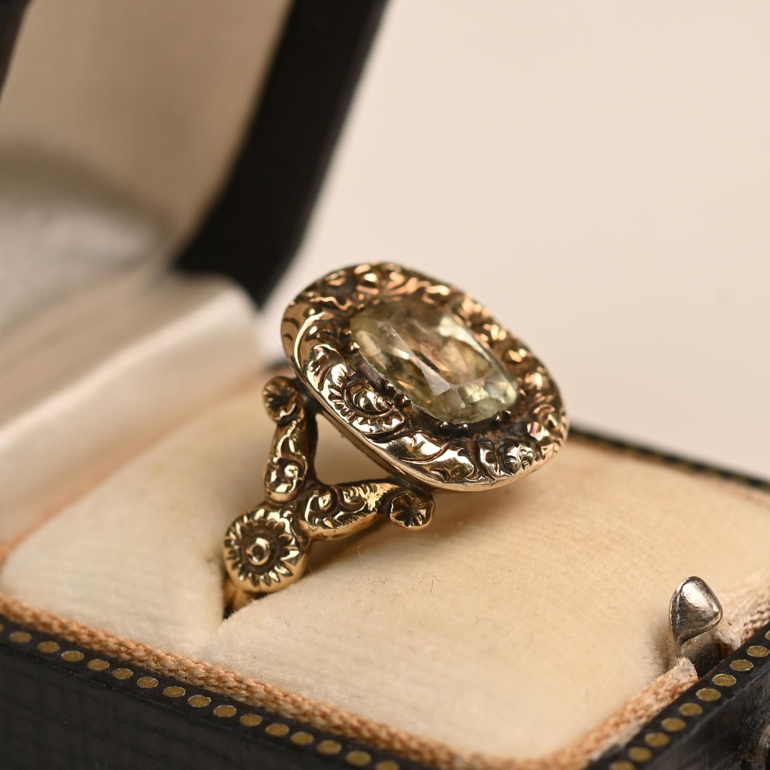 Este anillo, un raro tesoro de la época georgiana, exhibe un cautivador topacio talla cojín como pieza central, sujeto con seguridad en un clásico engaste de garras. Alrededor de la vibrante gema hay intrincados detalles grabados a mano que fluyen