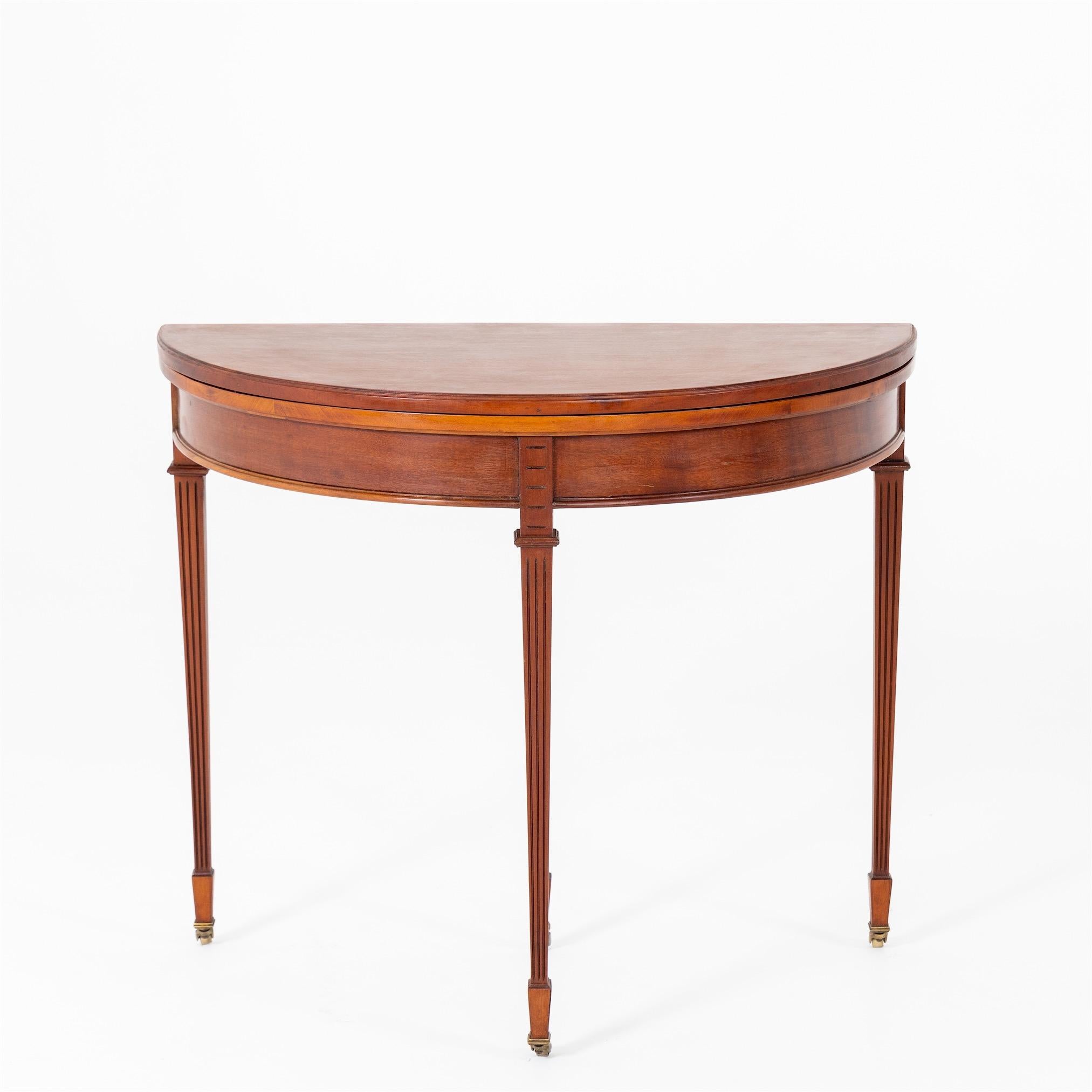 Demi-lune table made of mahogany, solid and veneered, and standing on four fluted pointed legs with brass castors. The hinged tabletop is covered with green leather on the inside.