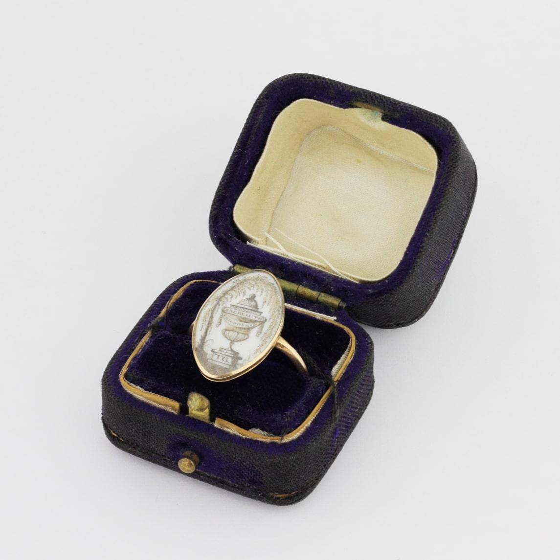 A George II mourning ring in its original box dating to 1781. To the rear is an inscription indicating it was in remembrance of Thomas Grimsby, a young sea captain. To the front is a hand painted urn with the initials 'TG', under what is likely an