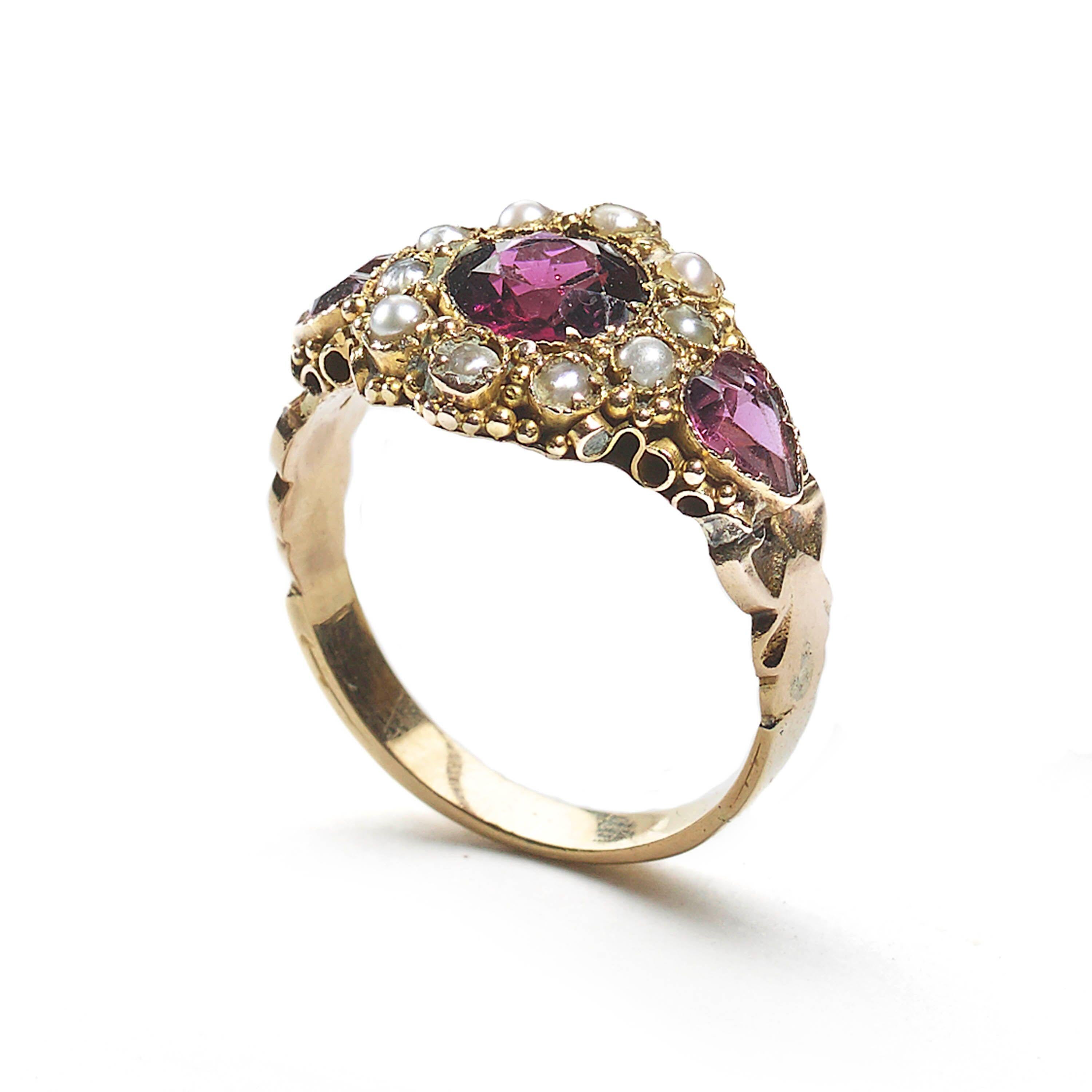 A Georgian almandine garnet and pearl ring, with a central round, faceted garnet, surrounded by ten, half seed pearls, with a heart-shaped garnet set in each shoulder, with granulated decoration, mounted in gold, with a chased  and engraved shank.