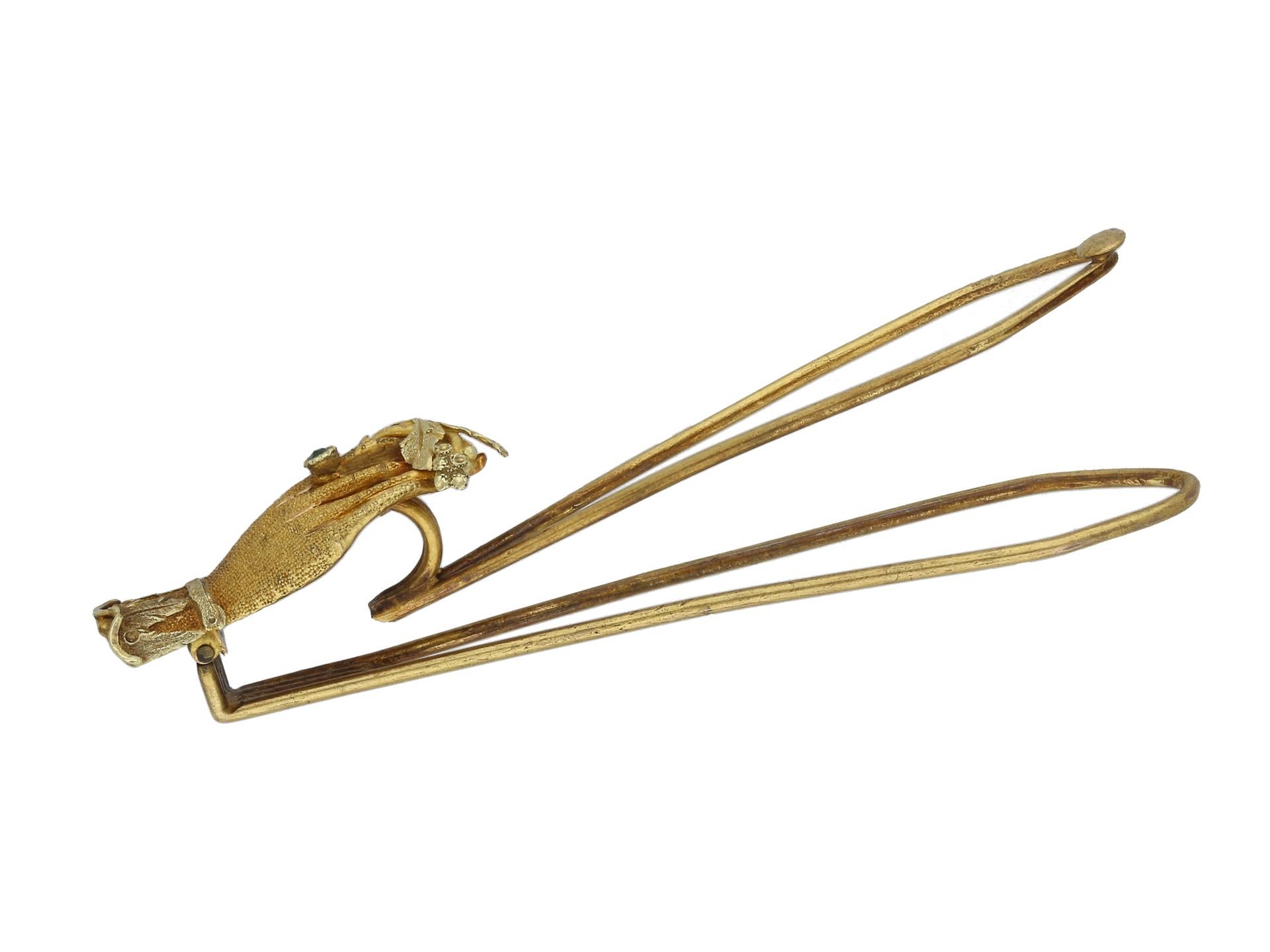 Georgische Haarspange oder Schalspange. Eine Haarspange aus Gelbgold mit einer Hand mit gekräuselter Manschette, die zwischen Zeige- und Mittelfinger einen Zweig mit Blättern und Beeren hält und einen Ring trägt, der mit einem kissenförmigen Smaragd