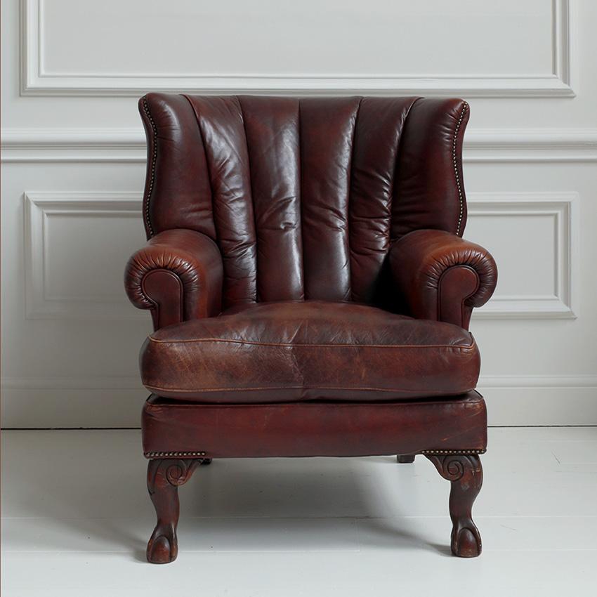 A Georgian-style wing armchair with rich, deep brown leather upholstery and worn patina, evoking a sense of heritage and timeless luxury. A signature Georgian detail, the nail head trim along the arms and edges adds a touch of refined craftsmanship