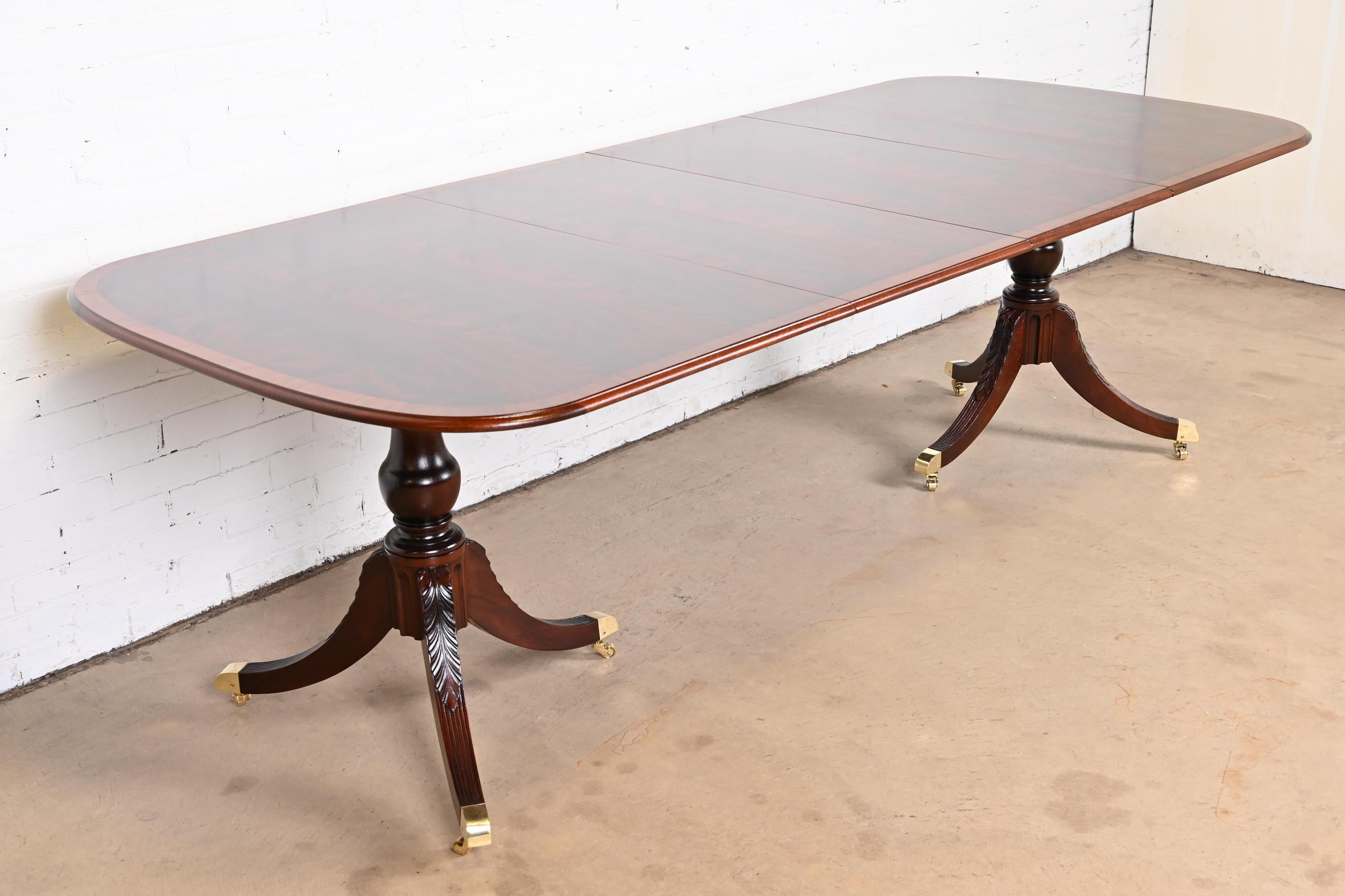 An exceptional Georgian or Regency style double pedestal extension dining table.

In the manner of Baker Furniture

USA, Mid-20th Century

Gorgeous book-matched flame mahogany, with satinwood banding, carved solid mahogany pedestals, and