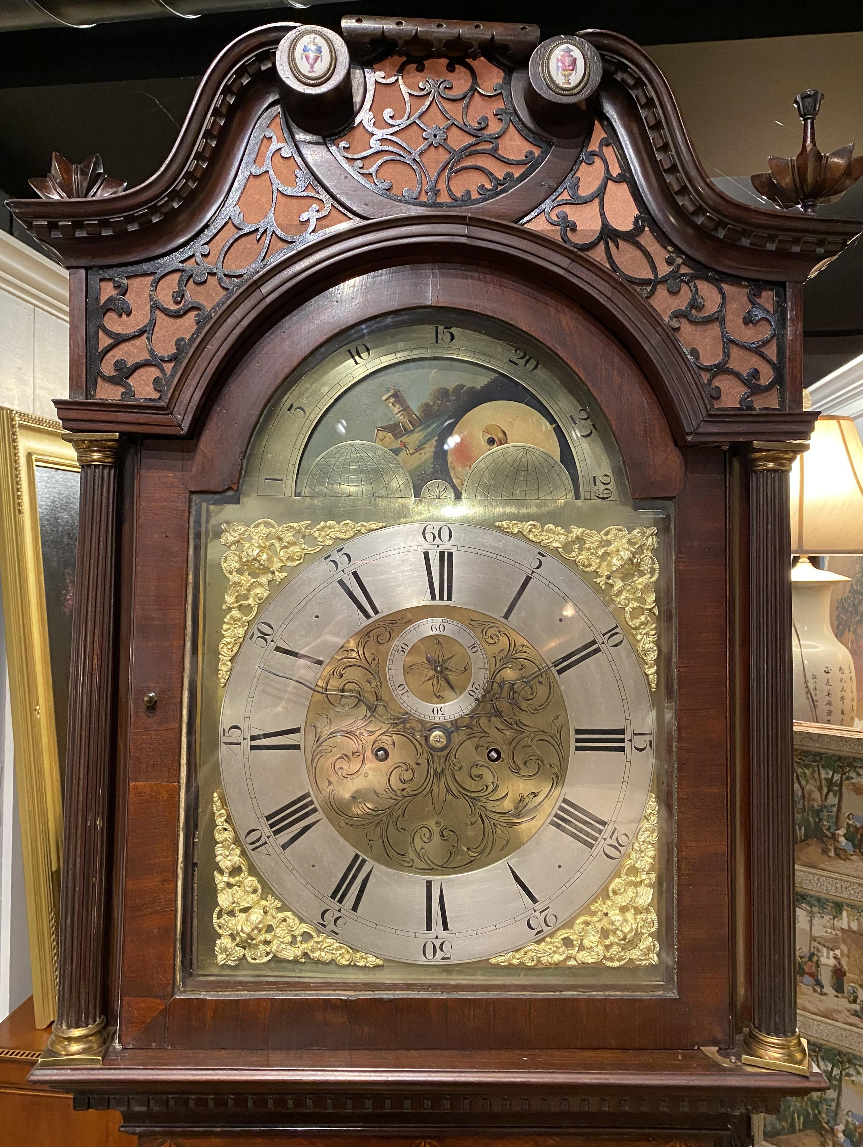 A fine English Georgian Gothic style tall case clock, its bonnet with a swan’s neck broken arch pediment with Battersea enamel oval polychrome urn accents surmounting scrolled fabric lined fretwork, flanked by two decorative carved finials, over a