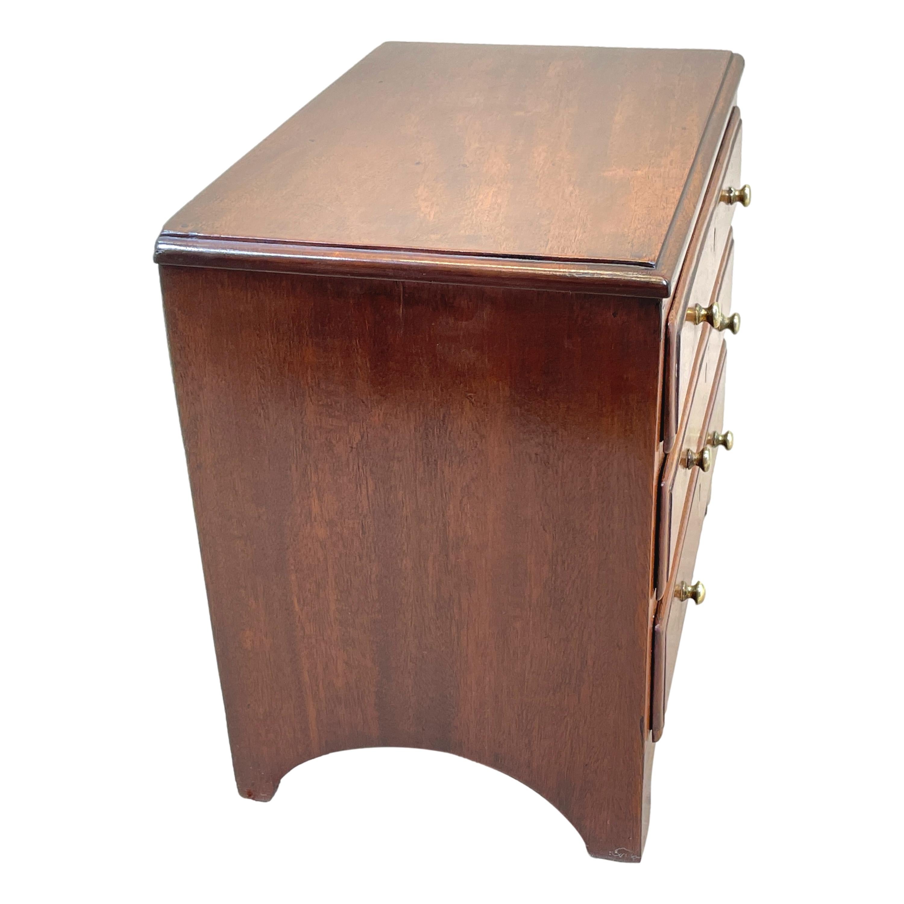 A Good Quality And Charming Early 19th Century Mahogany Miniature Chest, Having Well Figured Top Over Three Long Drawers With Original Brass Knobs And Boxwood Inlaid Escutcheons, Over Elegant Shaped Apron With Original Splayed Bracket