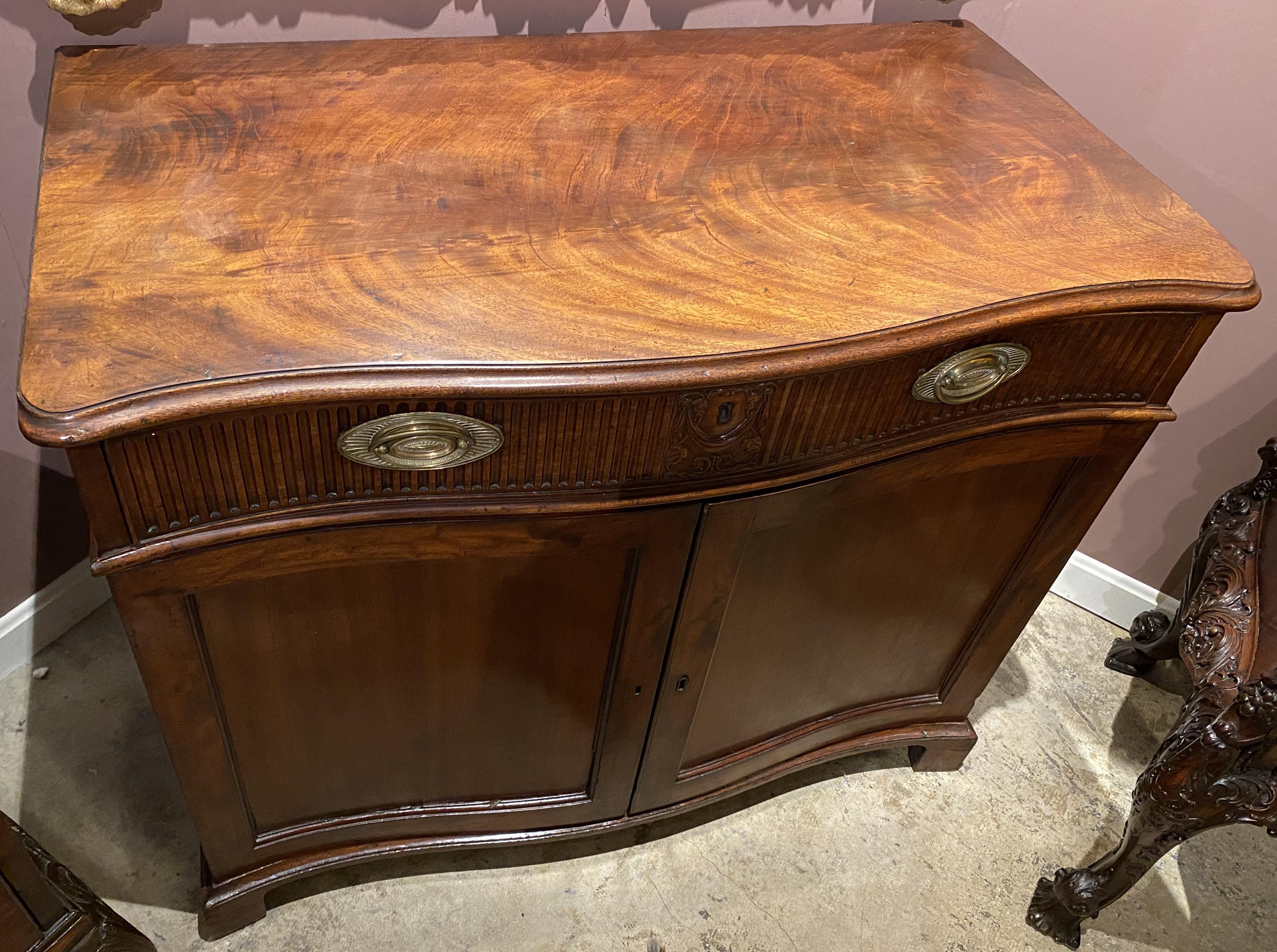 A fine hand carved Georgian mahogany butler’s desk with conforming molded edge top surmounting a slide out top drawer with hinged writing surface and fitted drawers, over two paneled doors which open to two slide-out shelves, oval brass drawer pulls
