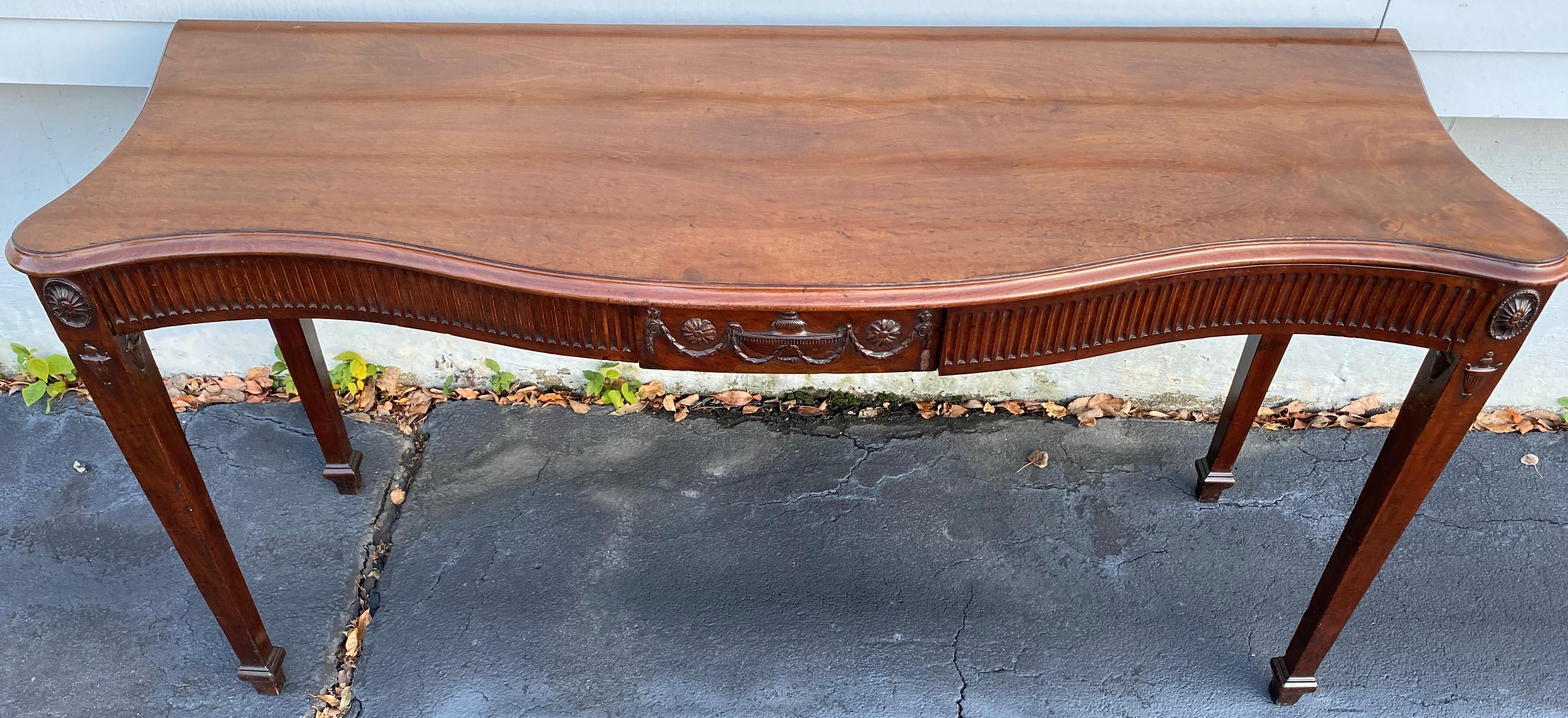 A fine Georgian mahogany serpentine sideboard, serving or console table with shaped molded edge top surmounting a frieze with central urn & swag carved panel flanked by two conforming reeded drawers, carved oval rosettes and urns on each corner,