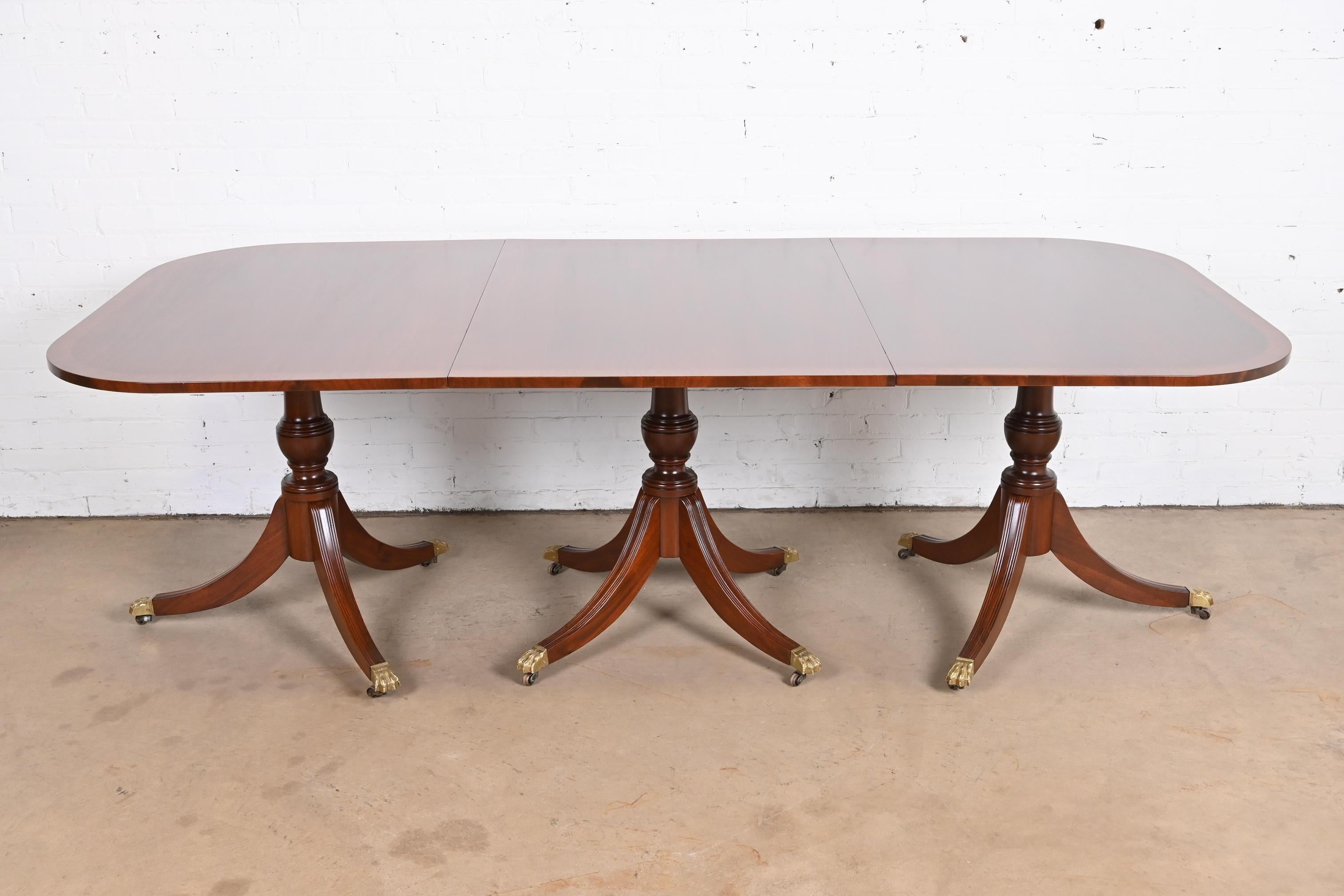 An exceptional Georgian or Regency style extension triple pedestal banquet or dining table.

In the manner of Baker Furniture.

USA, circa mid-20th century.

Mahogany, with satinwood banding, carved solid mahogany pedestals, and brass paw feet