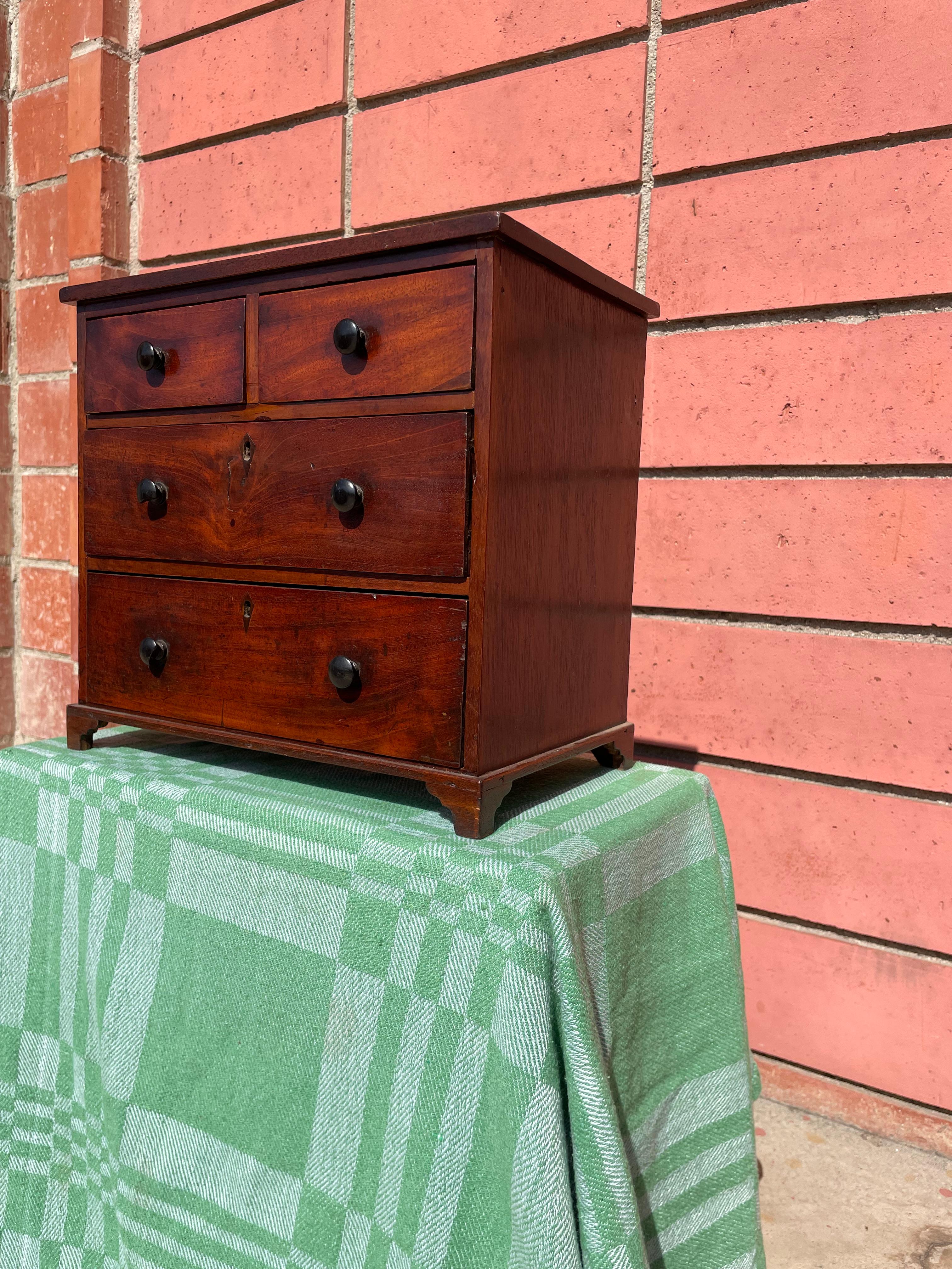 This early 19th century Georgian Miniature Chest of Drawers would have been constructed by the workshop to show potential clients examples of their products. Made from sturdy mahogany wood, raised on small detailed braket feet, hand carved-wooden