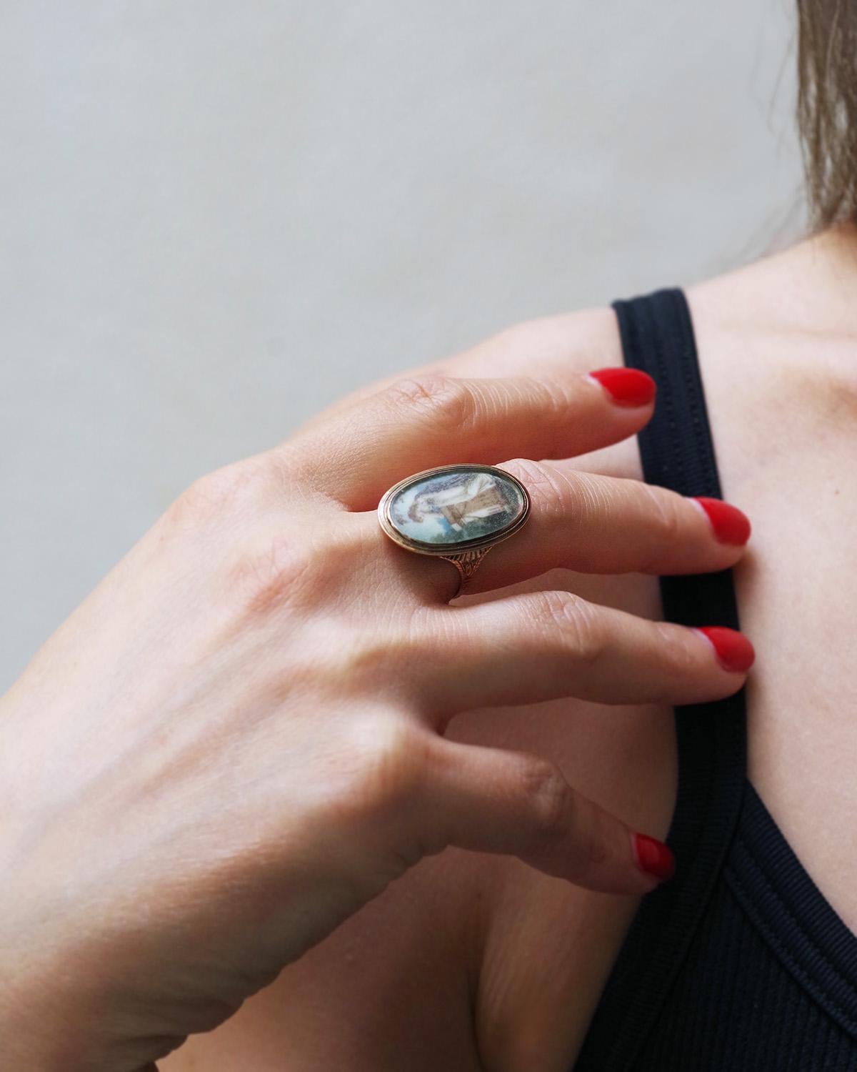 Georgian miniature mourning ring in low-grade gold. Oval in shape, this ring features a polychrome miniature in its center, protected by glass. The miniature depicts a young woman leaning on a column, in mourning, staring at the ground. She wears a
