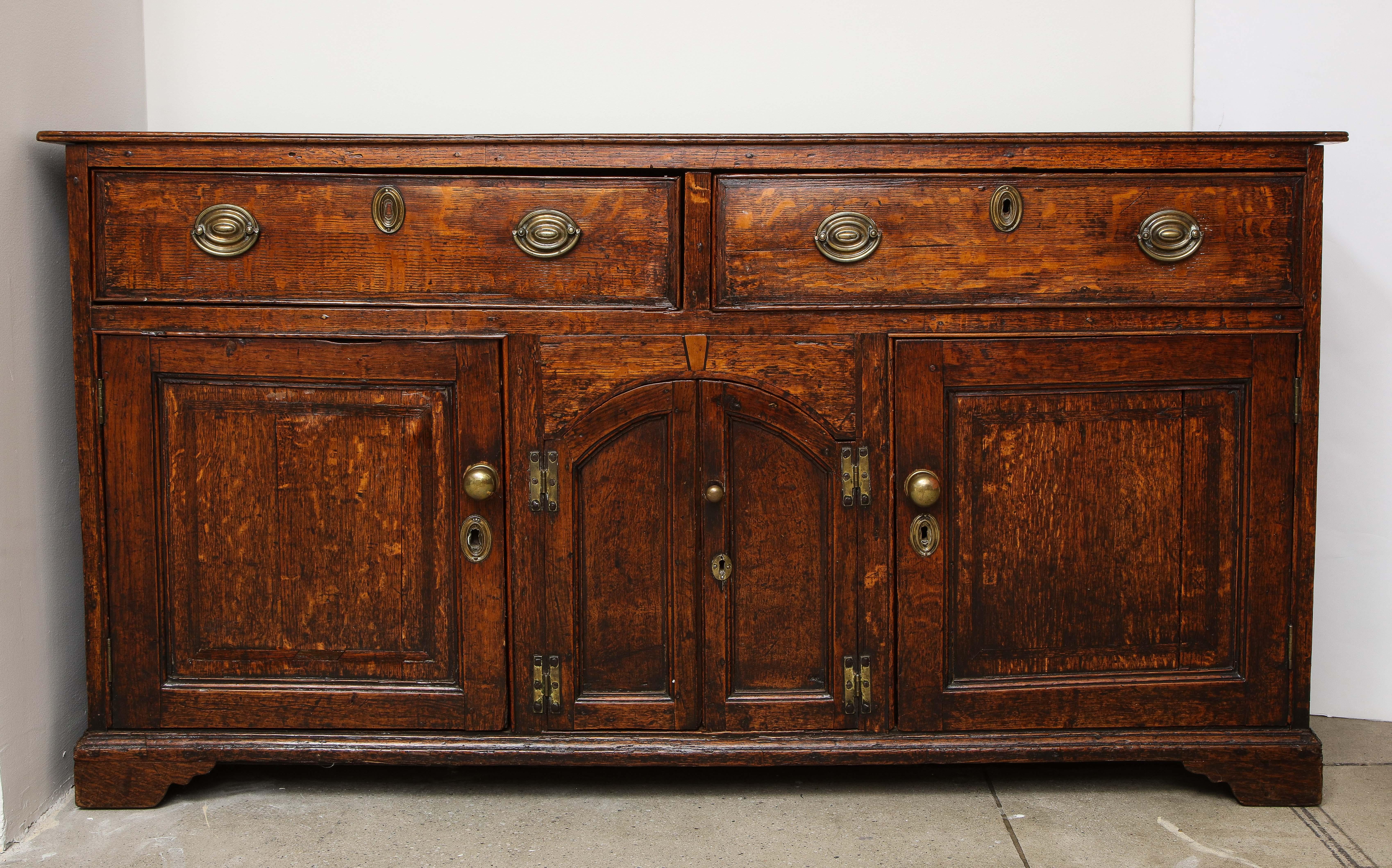 Fine George III oak low dresser of cabinet form, the top with rounded edge over two drawers with stamped brass pulls and escutcheons and mahogany cross banding, over pair of central arched panel doors retaining original brass 