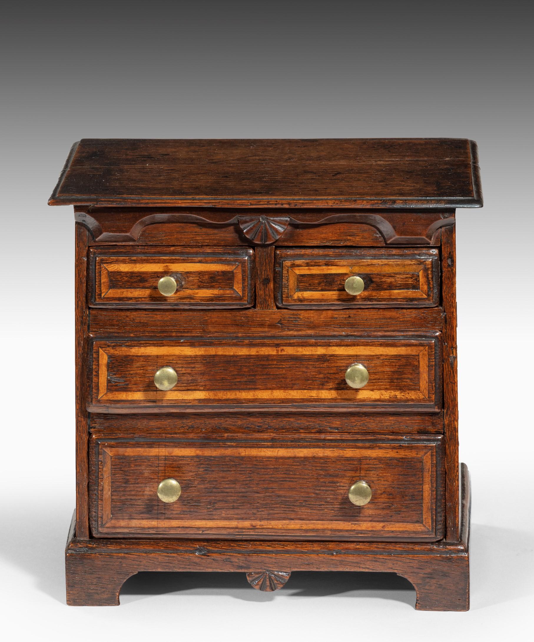 A charming George II oak miniature chest of drawers; the top above a shaped frieze centred by a scallop shell above two short and two long drawers which have ovolo mouldings and are inlaid with boxwood stringing; the miniature chest is raised on its