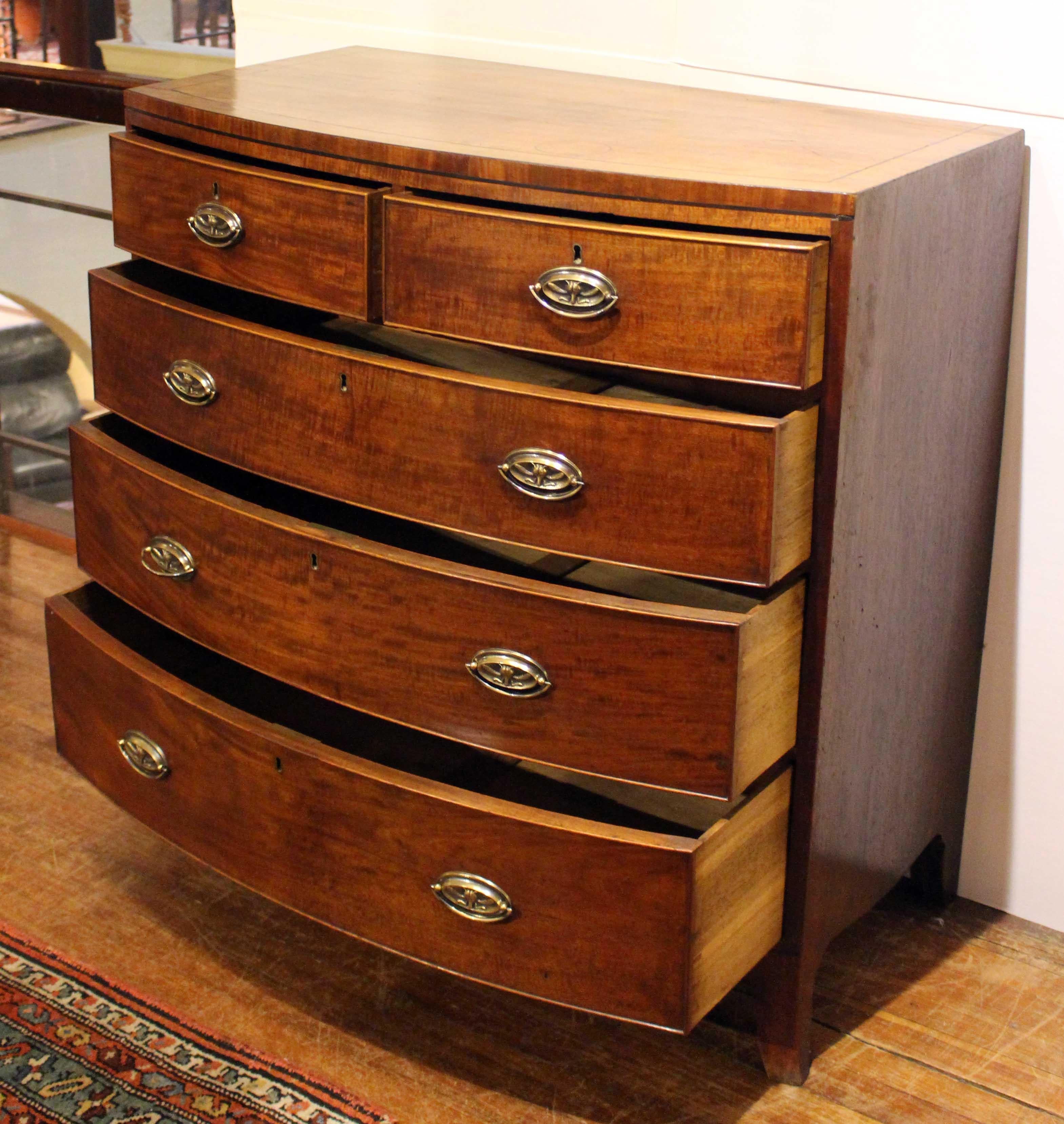 A Georgian period, c.1810, bowfront chest of drawers comprised of 2 short over 3 long drawers. Mahogany with ebony stringing. Oak & pine secondary woods. Replaced oval brass handles. Raised on French splay feet. Shaped apron complements the form.