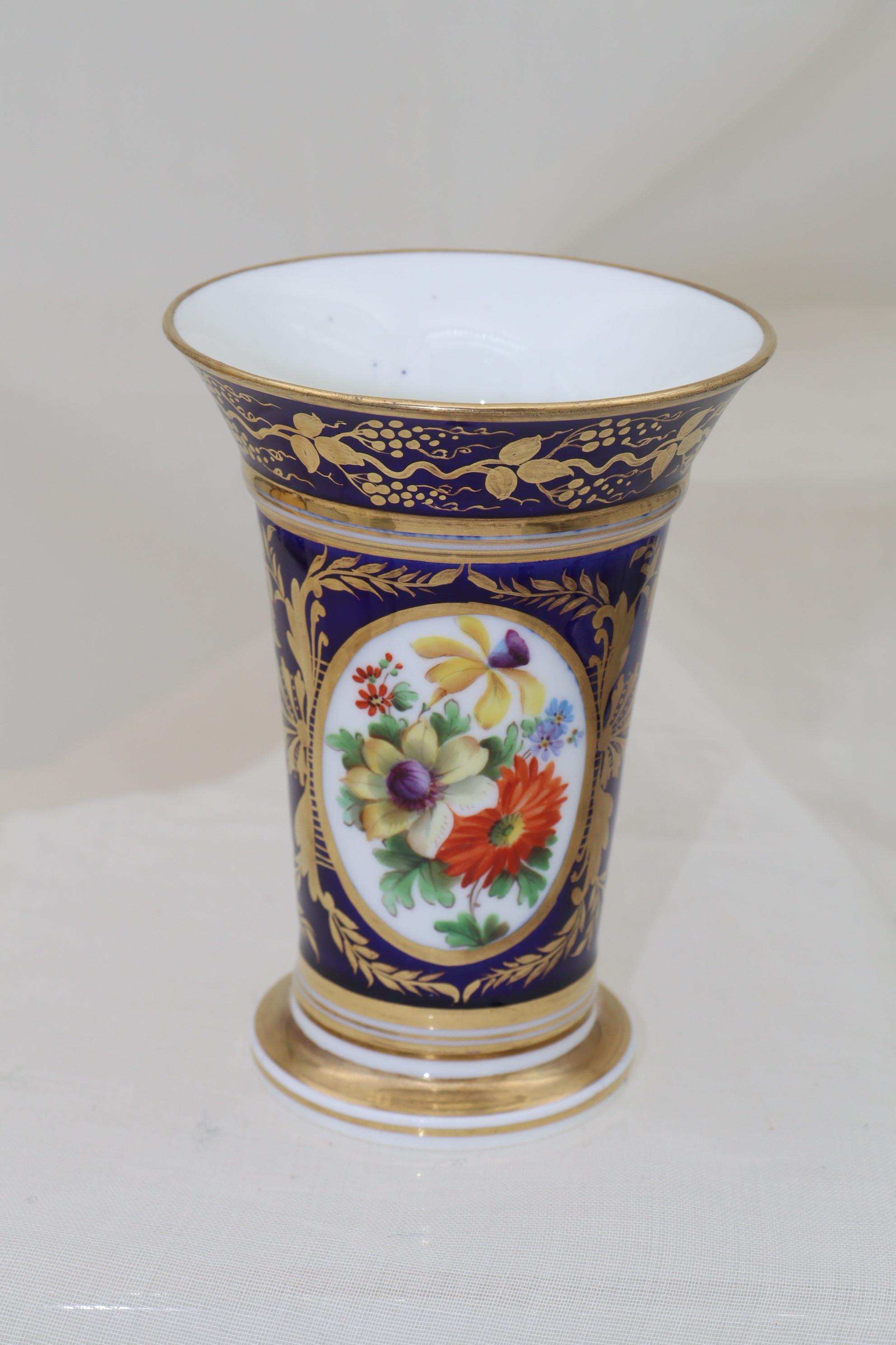 This Georgian hand painted and gilded porcelain spill vase features a gilded oval cartouche filled with a spray of colourful flowers. The cartouche is supported either side by an intricate gilded motif with more gilding to the rim and foot.