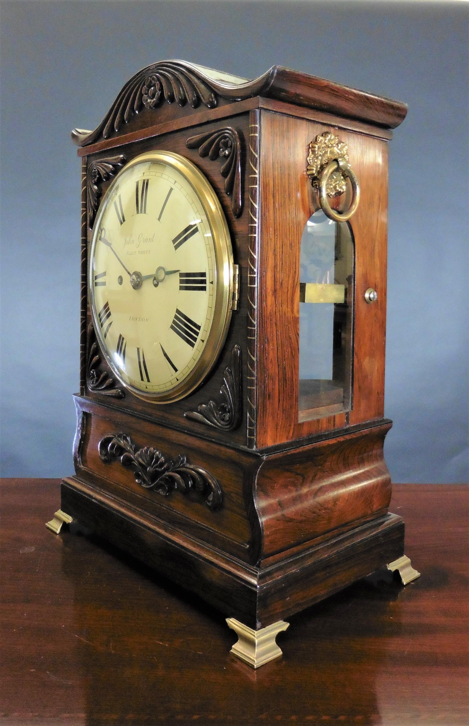 Georgian Bracket clock in a serpentine top rosewood case with applied carved decoration and side pilasters strung with brass inlay standing on brass claw bracket feet. 

Engraved cast bezel, original side fishscale frets and side handles. Convex