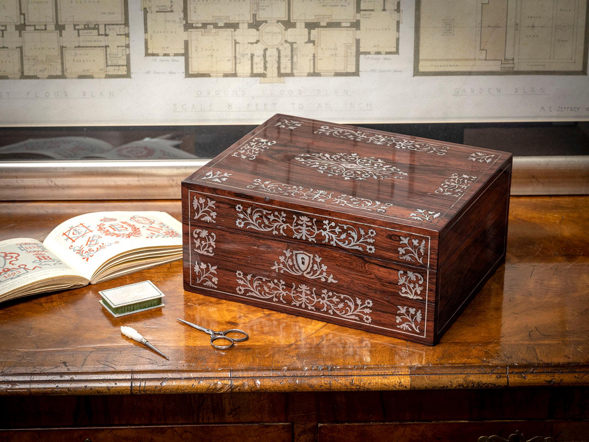 Aufwendige Perlmutt-Intarsien

Aus unserer Sewing Boxes Kollektion bieten wir Ihnen diese georgianische Palisander-Nähbox von Leuchars of London an. Der Nähkasten in rechteckiger Form ist aus Palisanderholz gefertigt und mit zwei von Silberschnüren