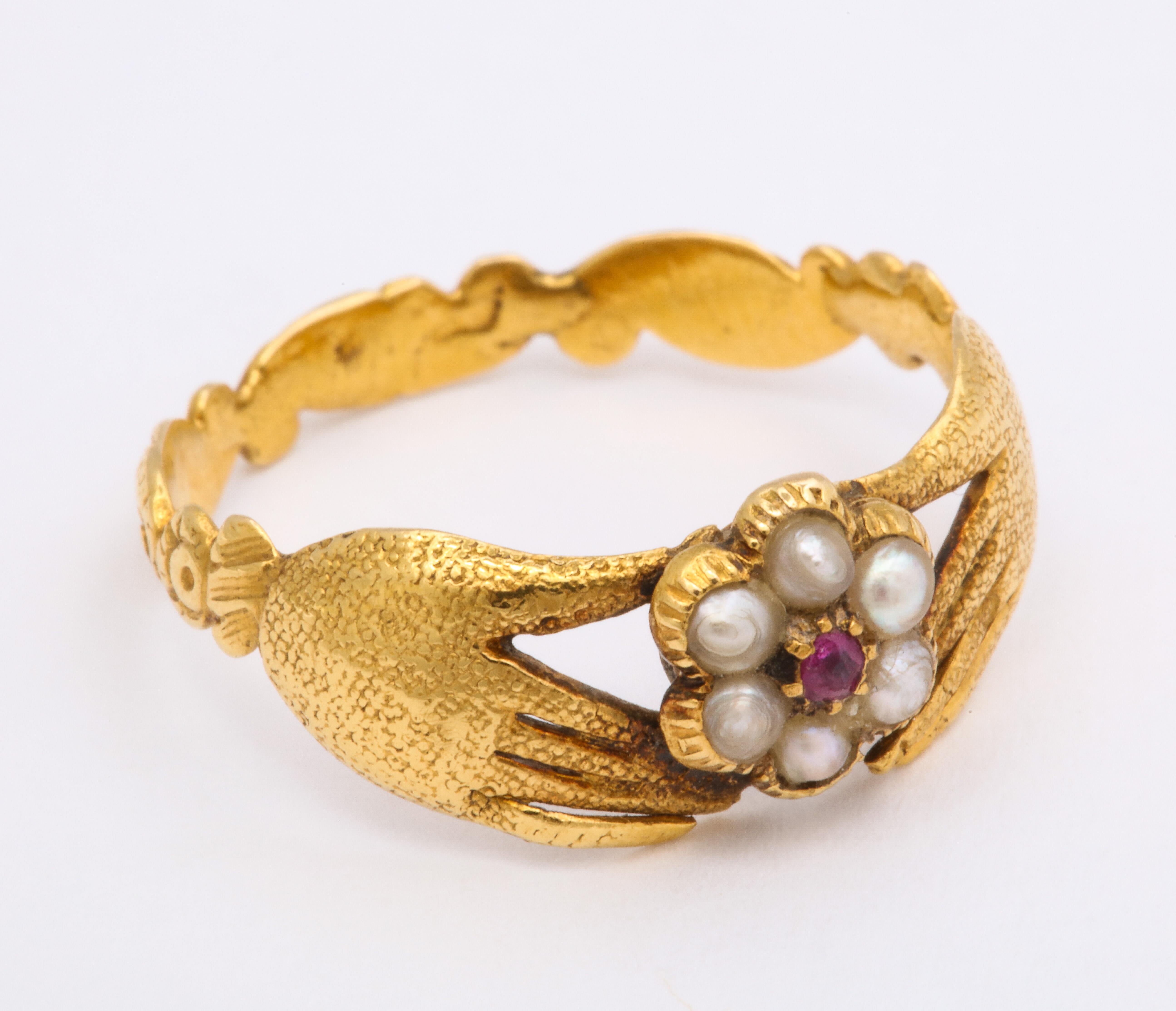 This English ring, made in 18K is a fede-like design. The hands themselves are not clasped but they clasp a central flower. The flower is made of a cluster of six pearls around a central ruby and has a locket back. Pretty engraved scroll shank.