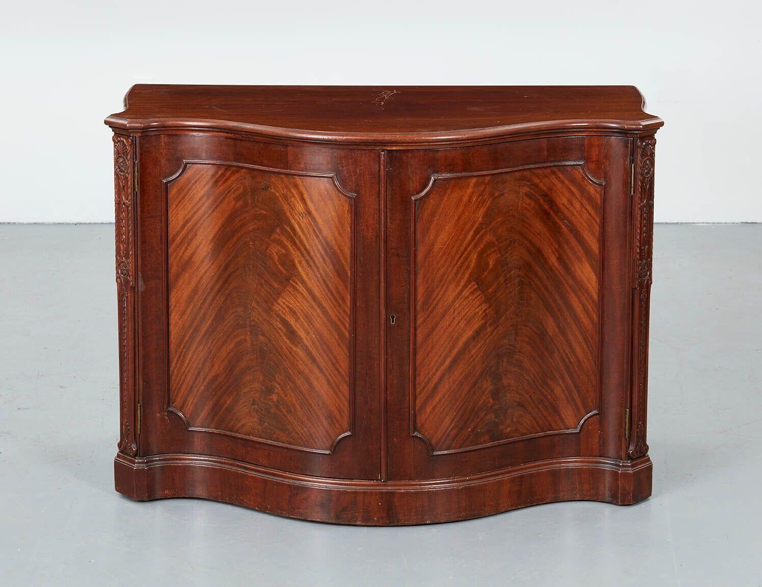 Very fine George III mahogany serpentine cabinet, the shaped top with molded edge, the two doors in vivid crotch grain with hard round molded surround, the canted corners having honeysuckle, bellflower and paterae carving and standing on molded