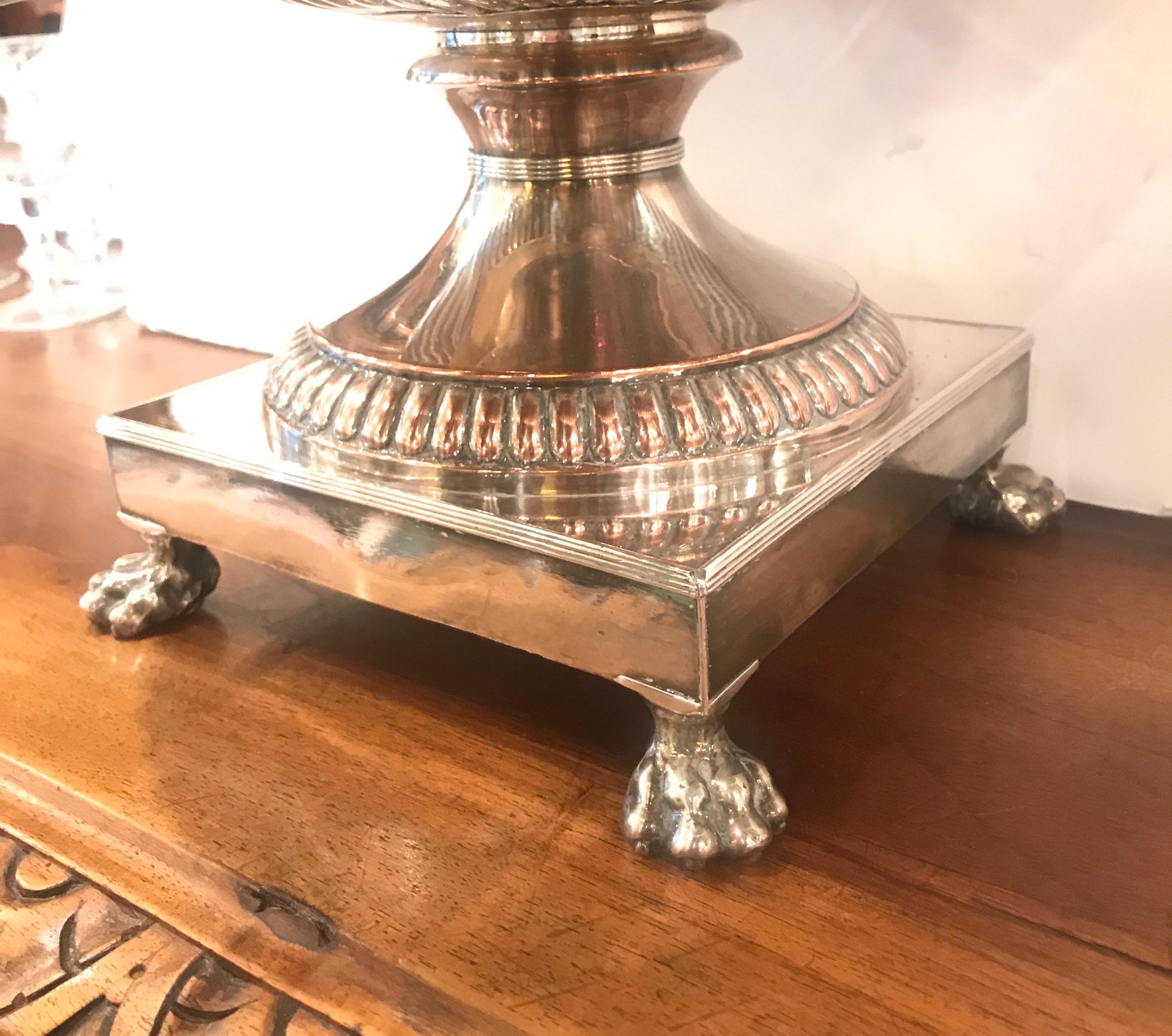 Of classical style, globular in design, the urn sits on a square pedestal base with four pawed feet, with two handles, mouthed spout. The reverted rim with heraldic device and the domed cover with leafed finial. This urn is accompanied with its