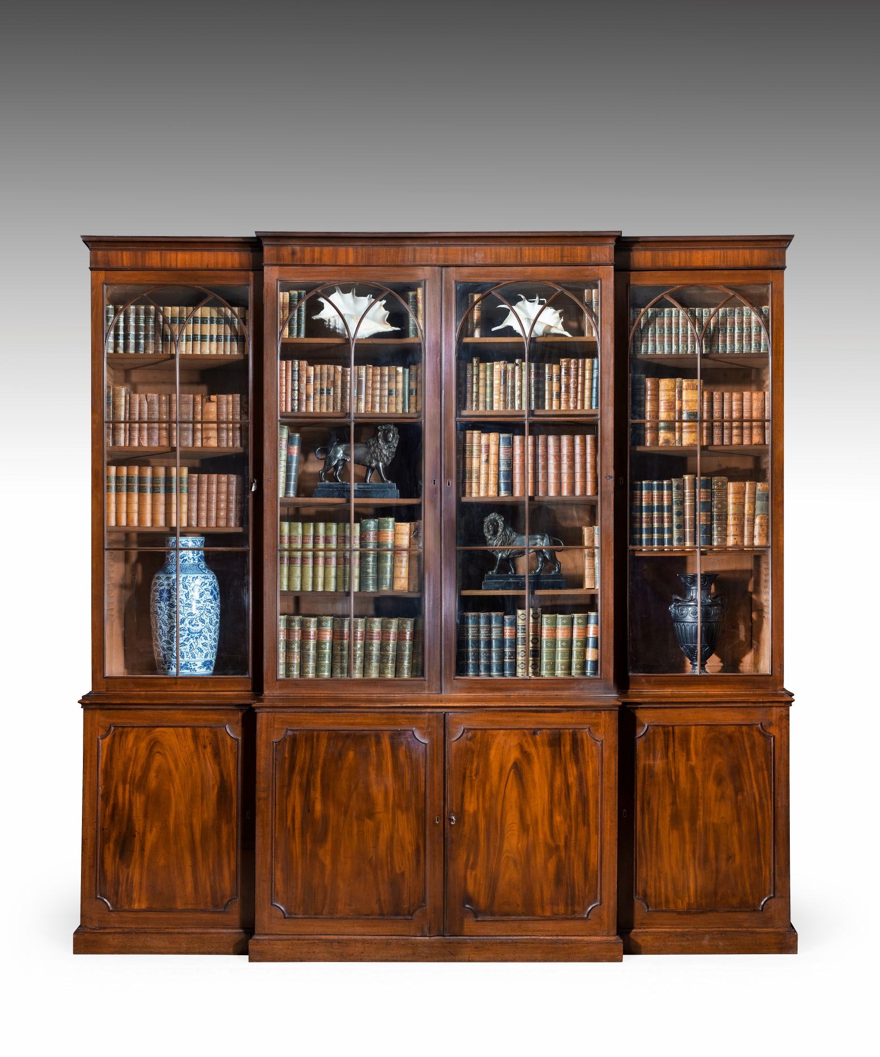 A George III Sheraton period mahogany breakfront bookcase; the cavetto moulded and crossgrain veneered cornice above glazed doors with elegant arched astragals that open to reveal fully adjustable shelves; below are panelled cupboard doors which