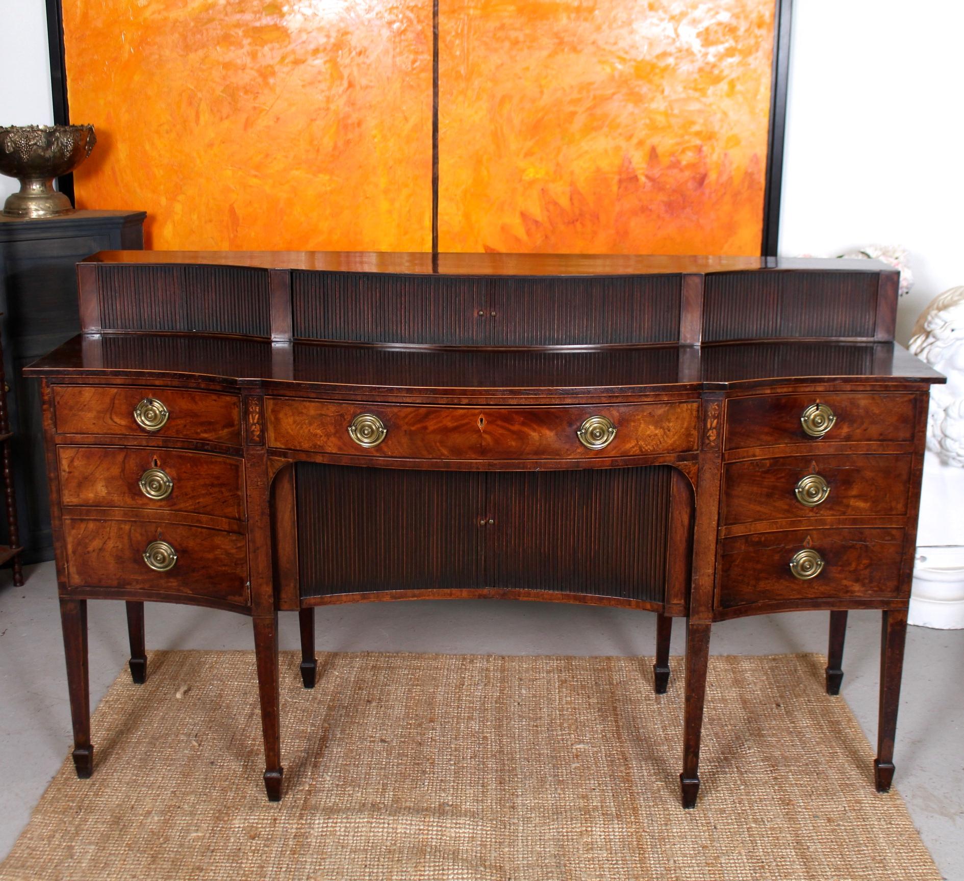 A fine quality rare form George III period serpentine sideboard.

The polished flamed mahogany inlays boasting an impressive grain.

The serpentine superstructure fitted central tambour doors and two outer tambour doors above a conforming