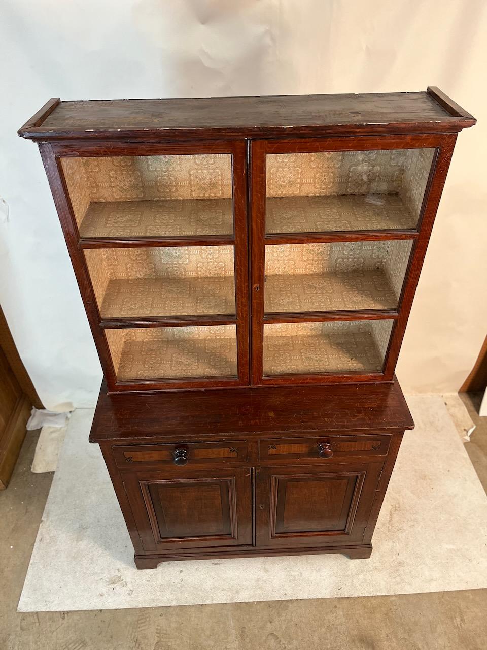 Classic Georgian step back 2 door book cabinet.  It has two drawers underneath with fleur-de-lys stenciling on the face of each drawer. Two doors below with one shelf storage.  Vintage wallpaper lining in the upper cabinet. Pine painted to resemble