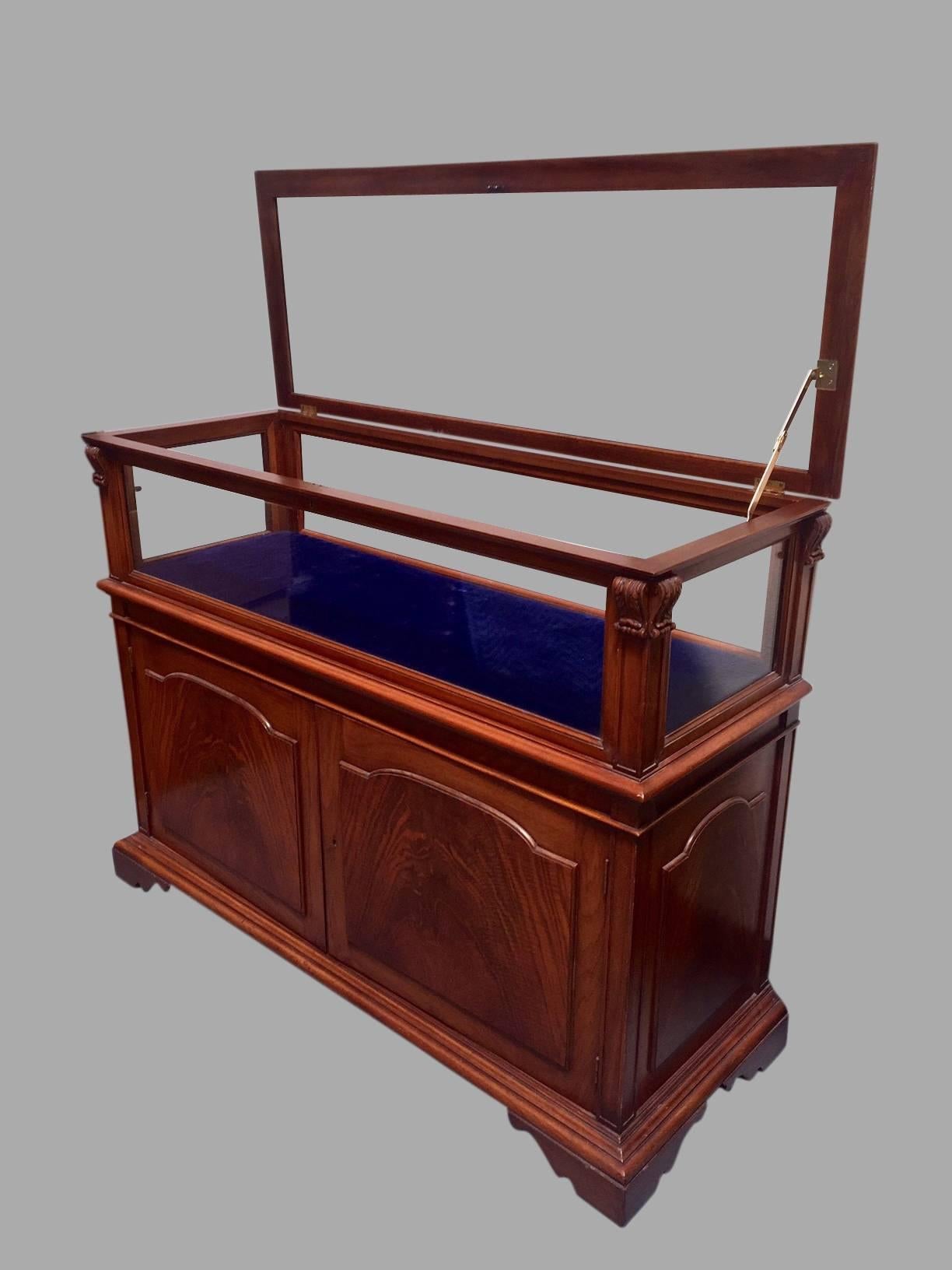 An attractive custom-made English or American mahogany showcase in one piece, the glazed upper section embellished with carved and fluted acanthus leaf decoration, the crotch mahogany veneered and paneled two-door lower section opening to reveal a