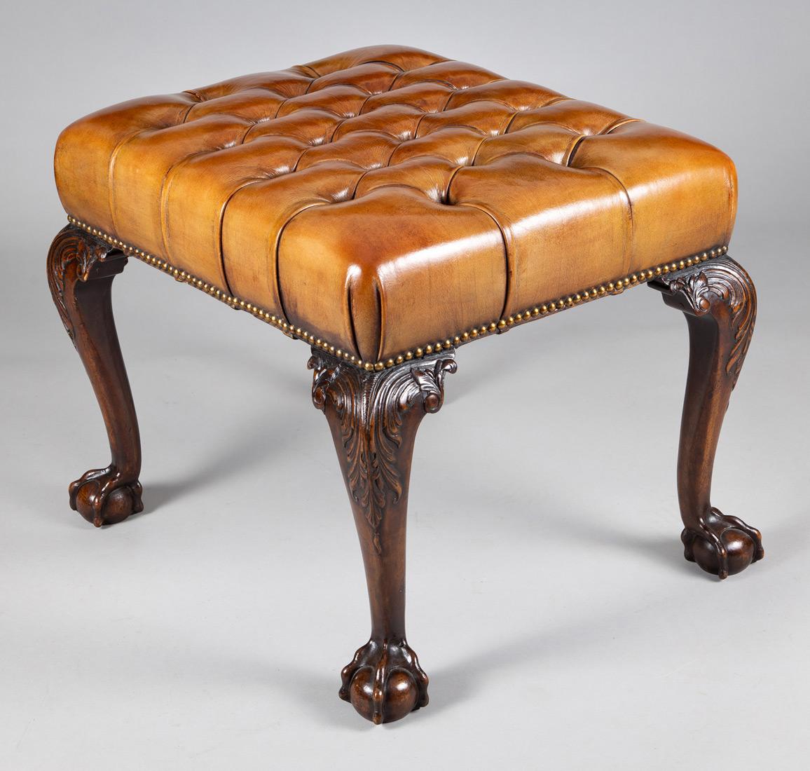 Georgian style mahogany stool upholstered in a cognac colored buttoned leather, supported on cabriole legs, the knees carved with acanthus leaves, ending in ball and claw feet. The leather is finished with brass nailheads.