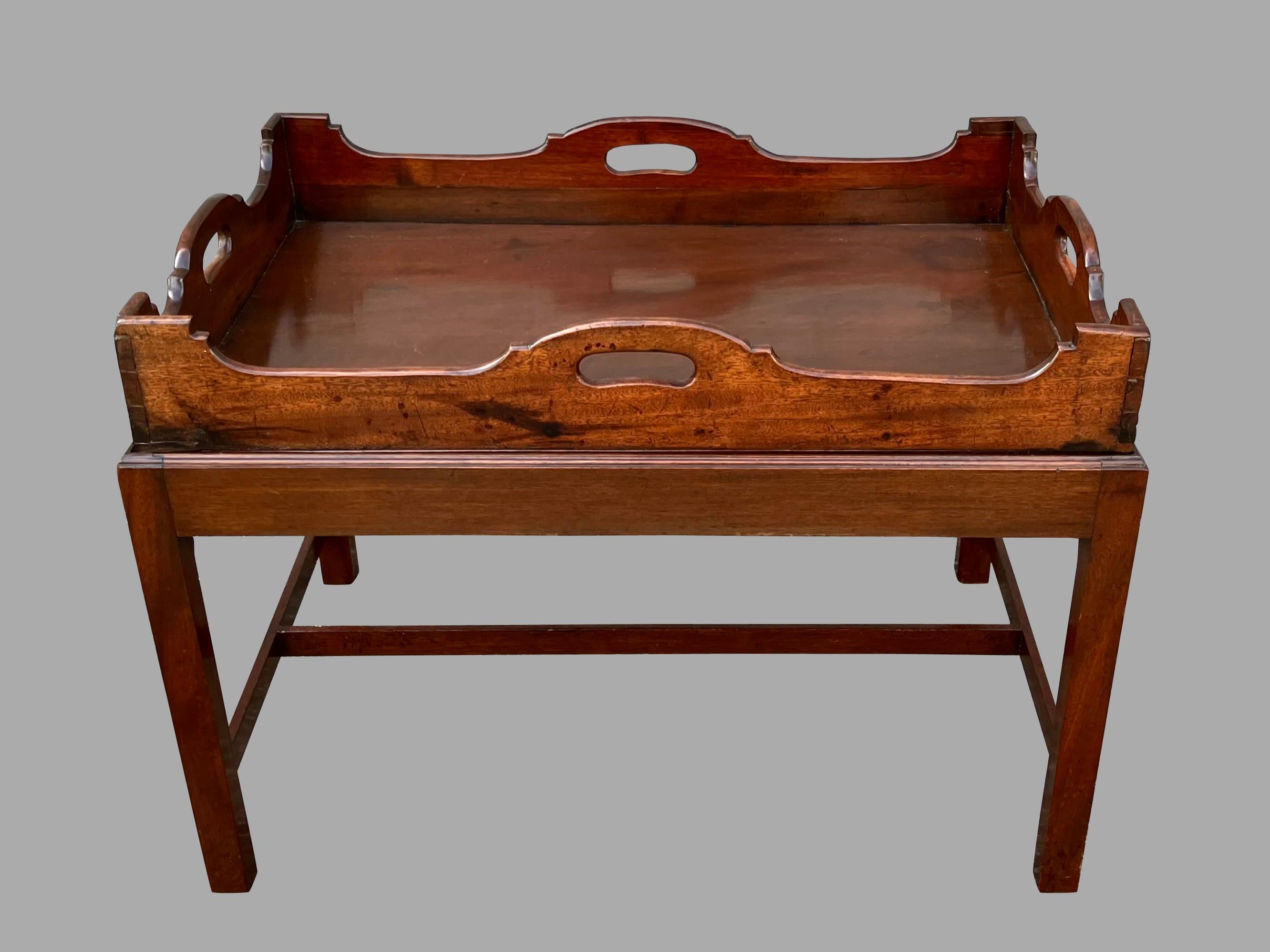 An English mahogany solid mahogany butlers tray with cutout side handles, the top edge of scalloped form, now resting on a custom Chippendale style stand of a later date with square legs joined by an H stretcher. The tray is removable from the stand
