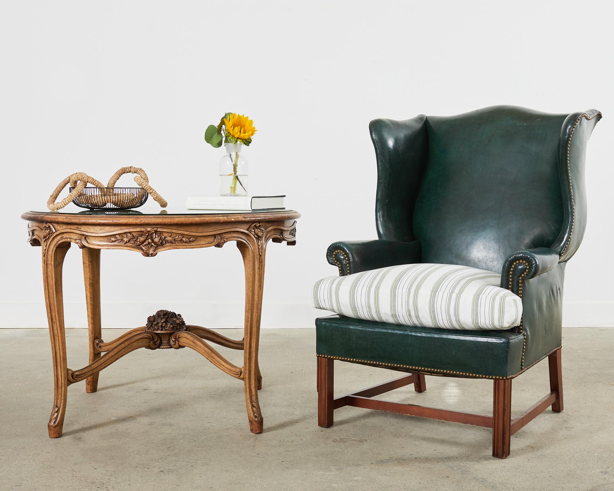 English Georgian style mahogany framed wingback chair featuring a regal hunter green leather upholstery. The large armchair features a hardwood mahogany frame with gracefully curved, fully developed wings. The frame has a classic hourglass shaped