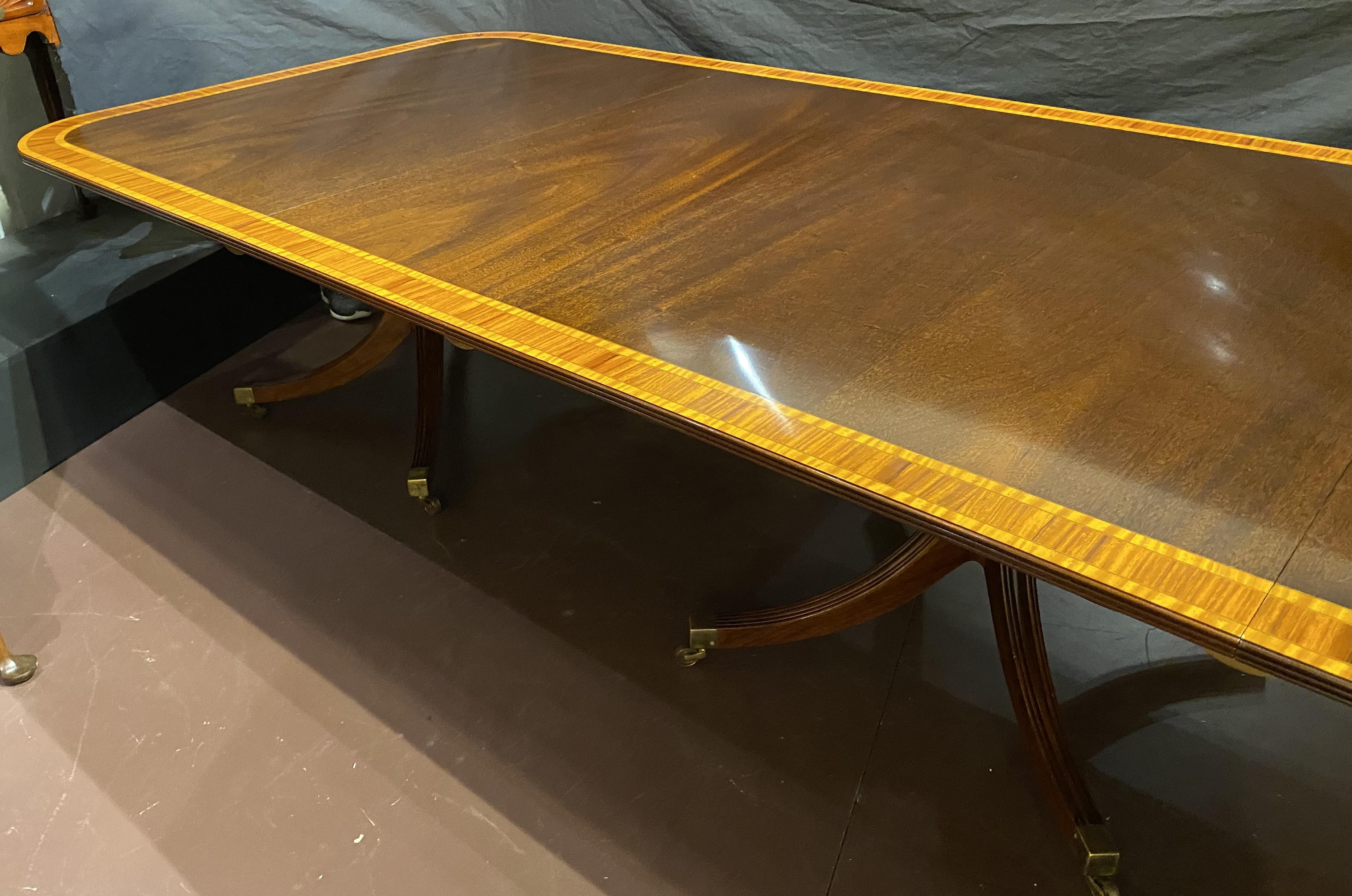 Table de salle à manger à trois pieds en acajou de style géorgien, avec des bandes de bois satiné et deux rallonges. Chacune des trois sections de la table est soutenue par quatre pieds cannelés et inclinés vers le bas, terminés par des roulettes à