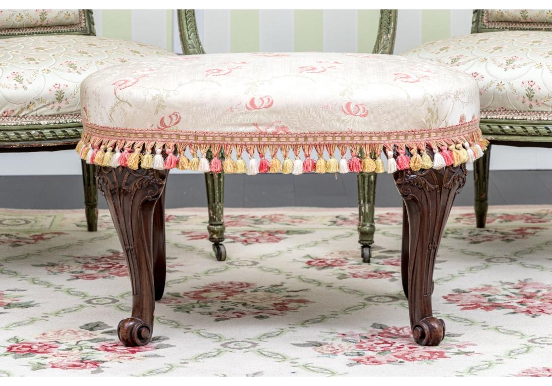 A finely constructed Georgian style oval mahogany bench raised on curved legs with wide knees with scrolled raised ribs ending in snail head turned feet. Upholstered in a fine pink floral printed ecru fabric with coordinating tassel trim.