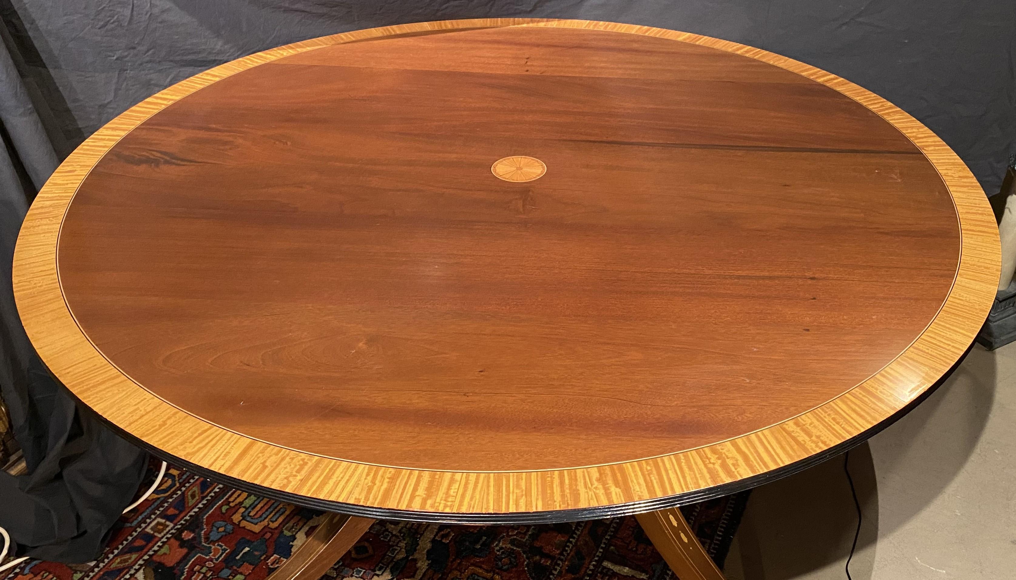 A fine example of a Georgian style round mahogany dining table with a satinwood banded veneer top with central pinwheel inlay, supported by a cage pedestal with turned supports and four outswept legs with line and bellflower inlay, terminating with