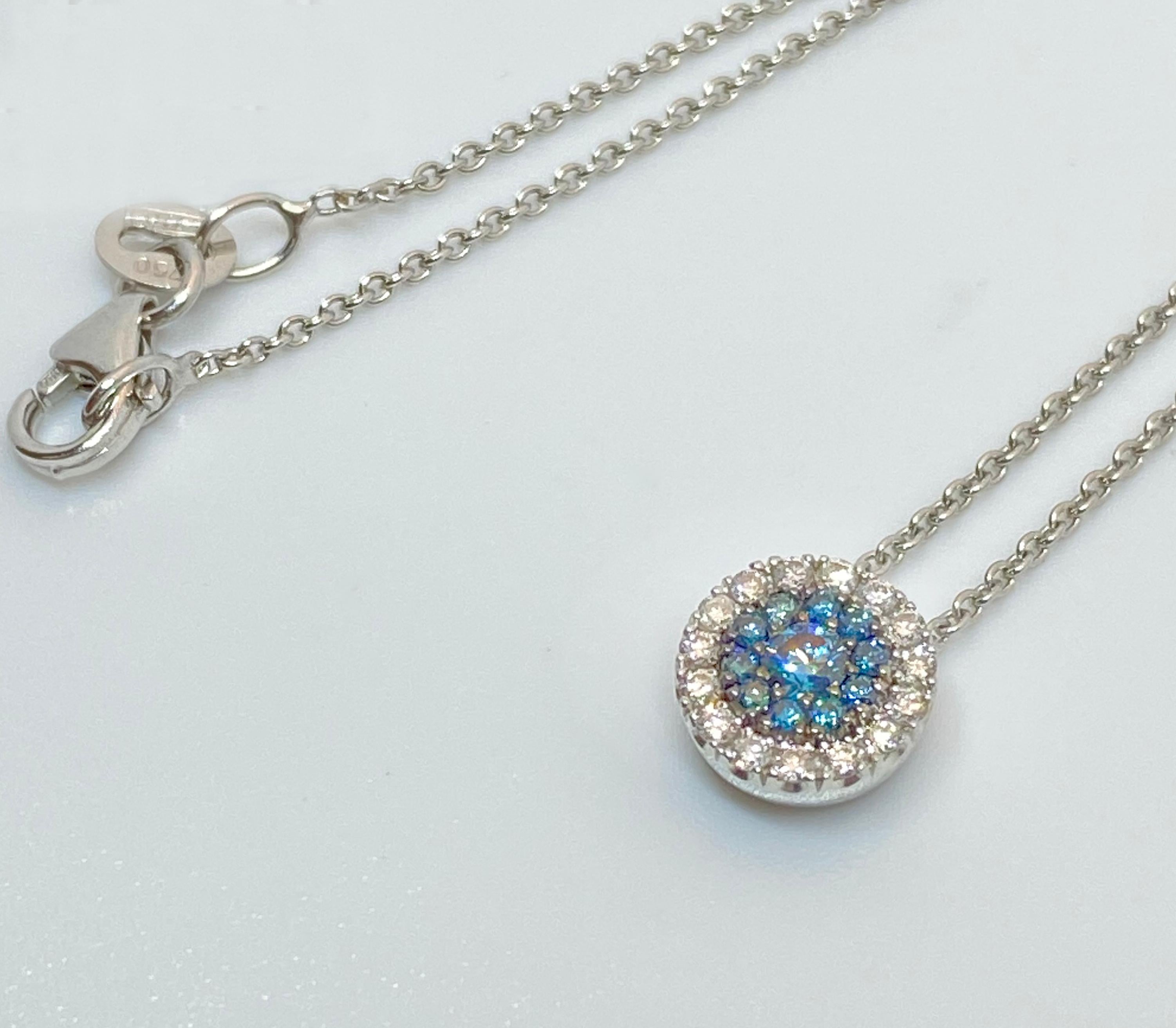 S.Georgios designer pendant Necklace is hand-made from 18 Karat White Gold to create a classic and elegant look. This beautiful Pendant Necklace feature a center brilliant-cut Blue Diamond with a total weight of 0.06 Carat, surrounded by 10 Blue