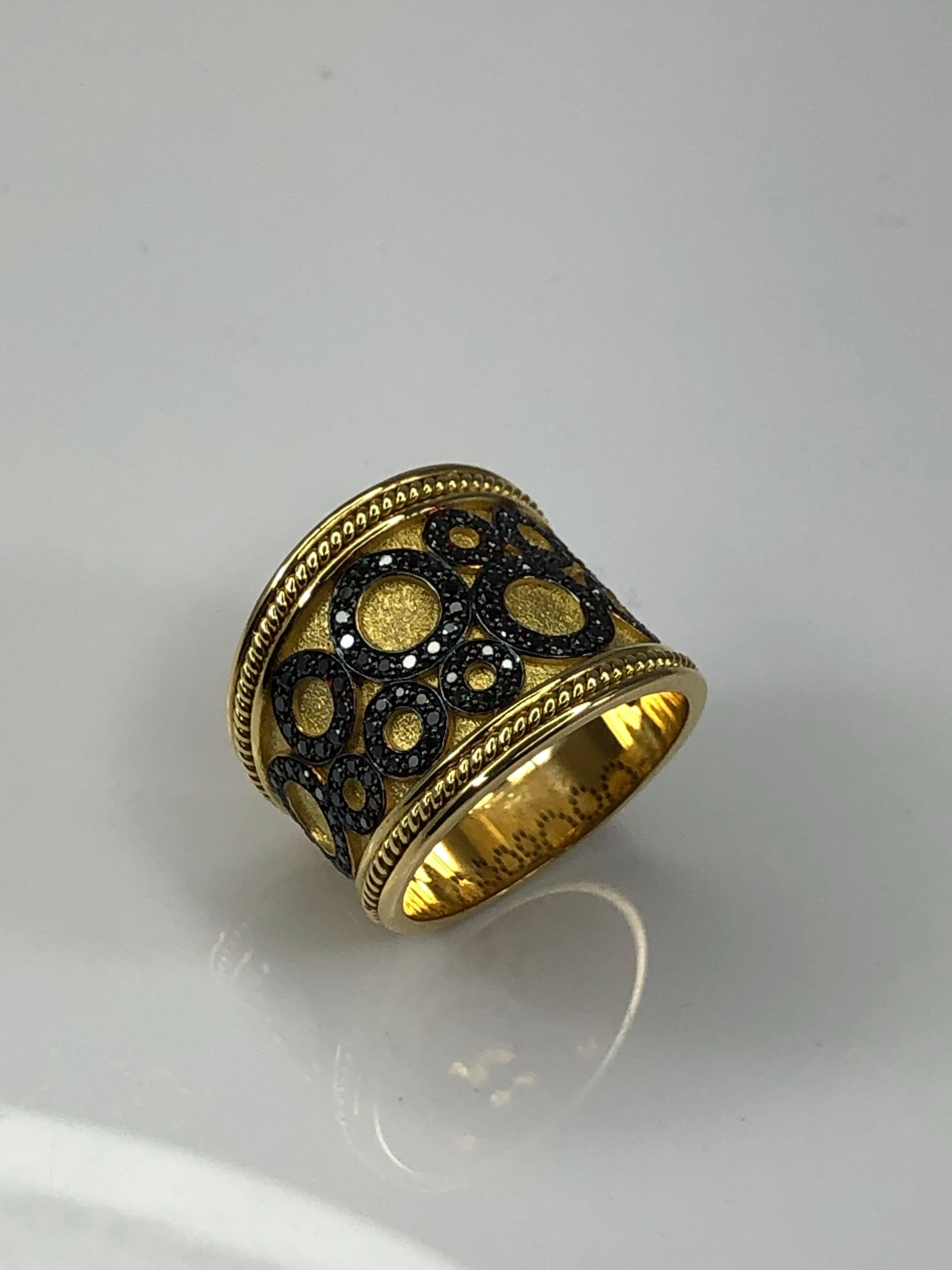 Ti presentiamo l'anello di design S.Georgios realizzato a mano in oro giallo 18 carati in Grecia. Questo anello combina l'antica tecnica bizantina con un design moderno. Su uno sfondo di velluto spiccano dei cerchi decorati con diamanti neri per un