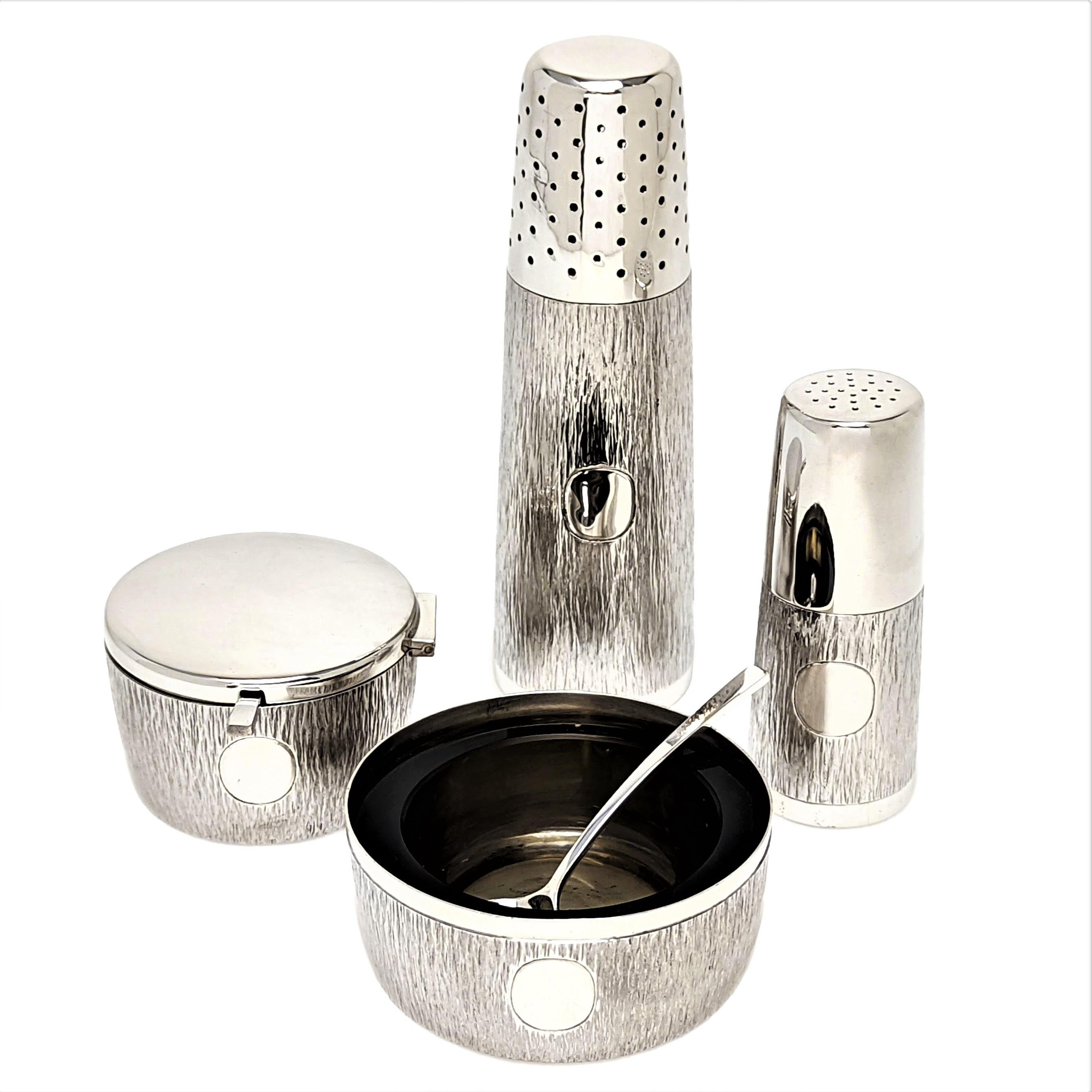 A modernist Gerald Benney Sterling Silver Condiment Set comprising of a Salt Pinch Pot, a Pepper Shaker, a Mustard Pot & a Caster. The Salt and Mustard have transparent brown glass liners and each has a silver spoon. The exterior of each item has an