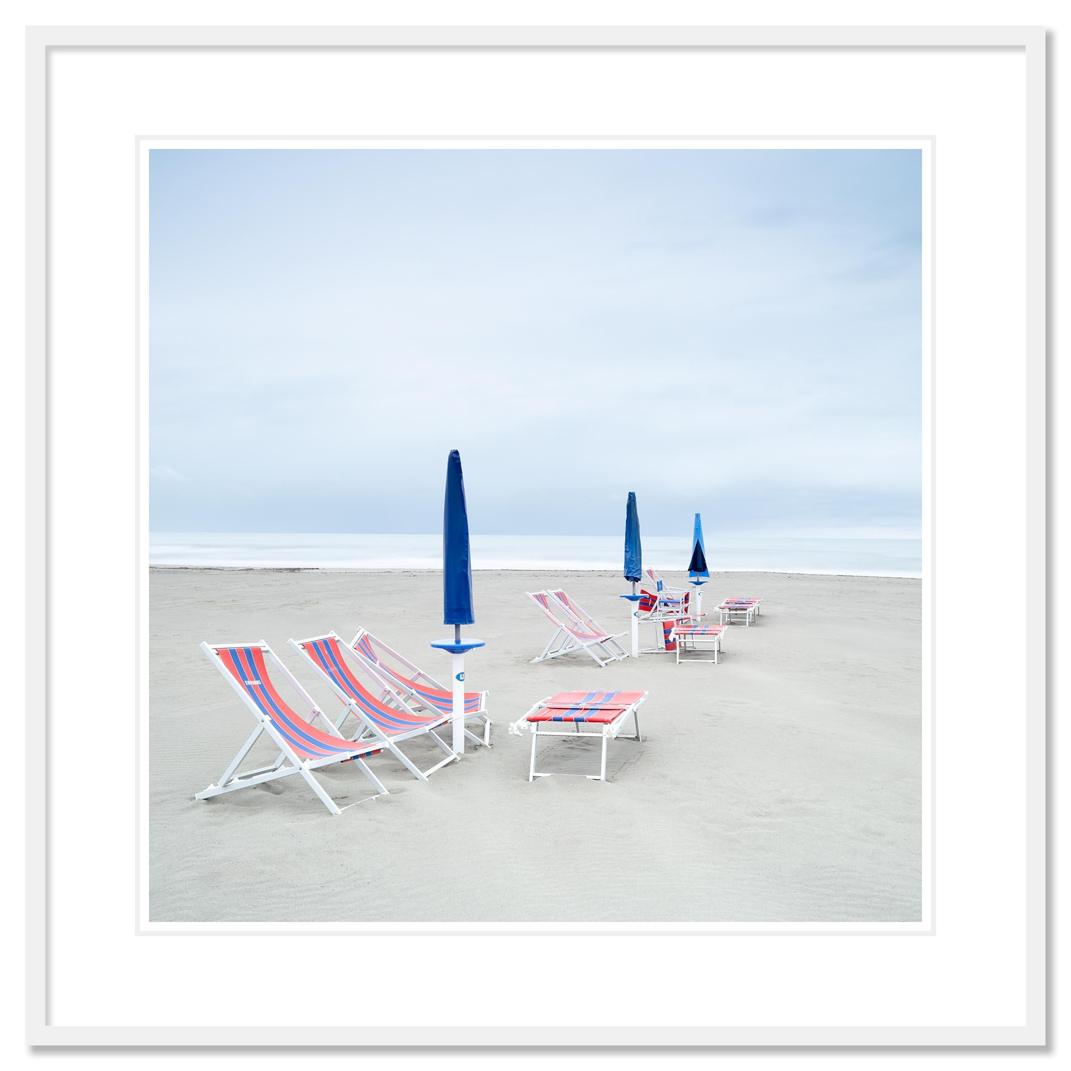 Gerald Berghammer - Photographie de paysage minimaliste en couleur. Plage vide avec des chaises longues rayées et des parasols bleus sur un rivage sablonneux, ciel couvert.

Tirage à l'encre pigmentaire d'archives faisant partie d'une édition
