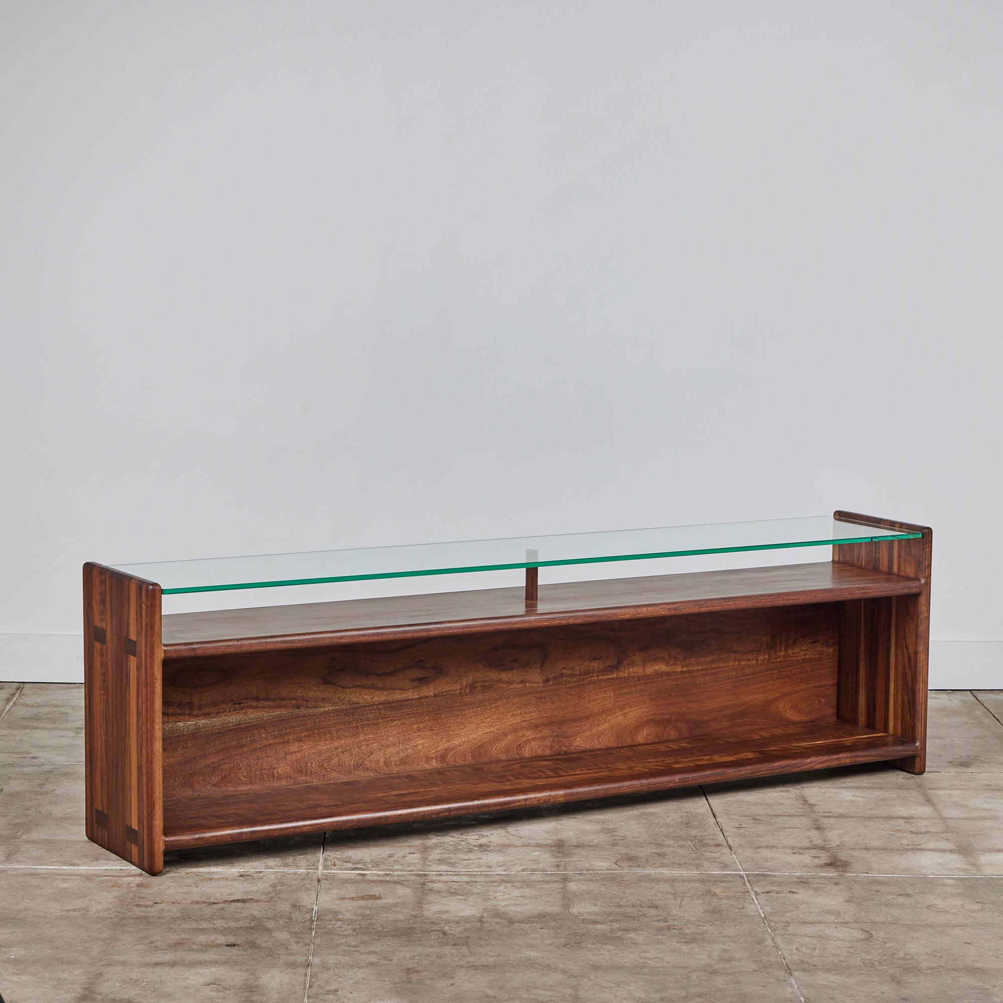 American made console table, circa 1970s, by Gerald McCabe is beautifully crafted from a solid slab of Shedua. The piece features natural wood grain variances with highs and lows. The top of this console showcases a glass top with a s shelf below