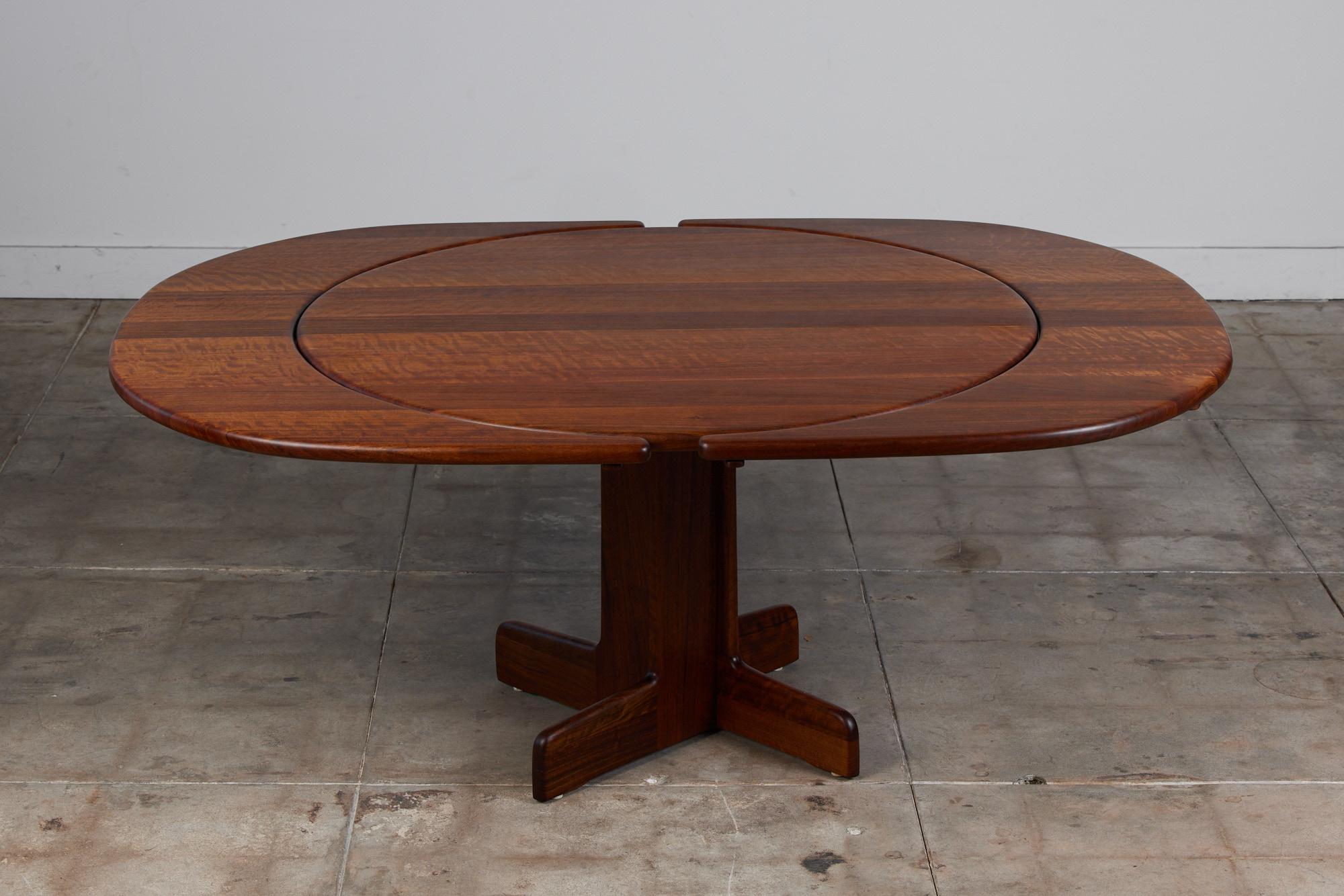American made dining table, c.1970s, by Gerald McCabe. The table features a round top situated on a pedestal base made of solid Shedua. The table comes with two crescent leaves which attach to the outers sides of the table expanding to seat six