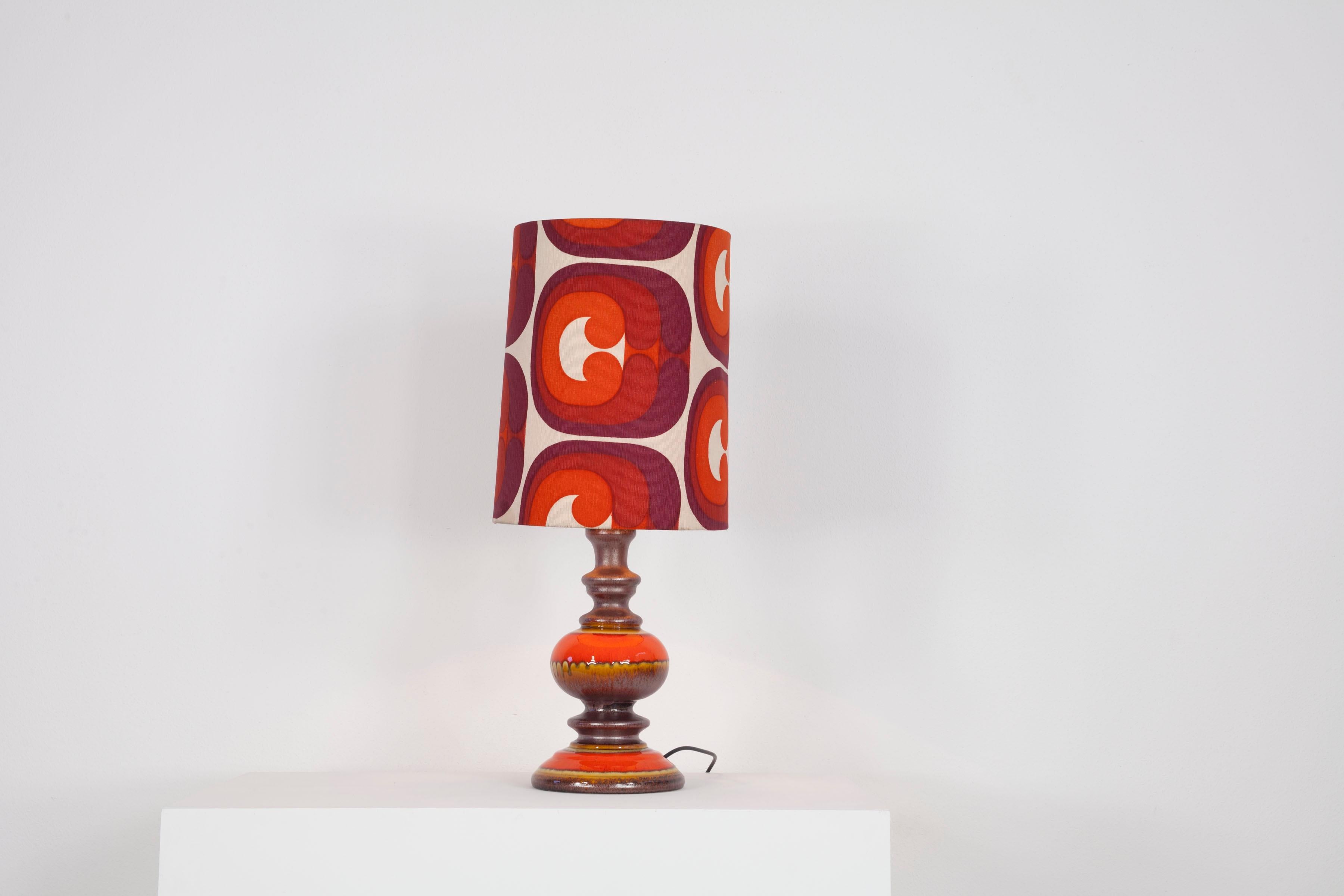 Découvrez le charme rétro des années 1970 allemandes avec cette lampe à poser en céramique aux tons chauds orange et marron. L'abat-jour spacieux en tissu complète le design et ajoute une atmosphère chaleureuse à n'importe quelle pièce. Une pièce