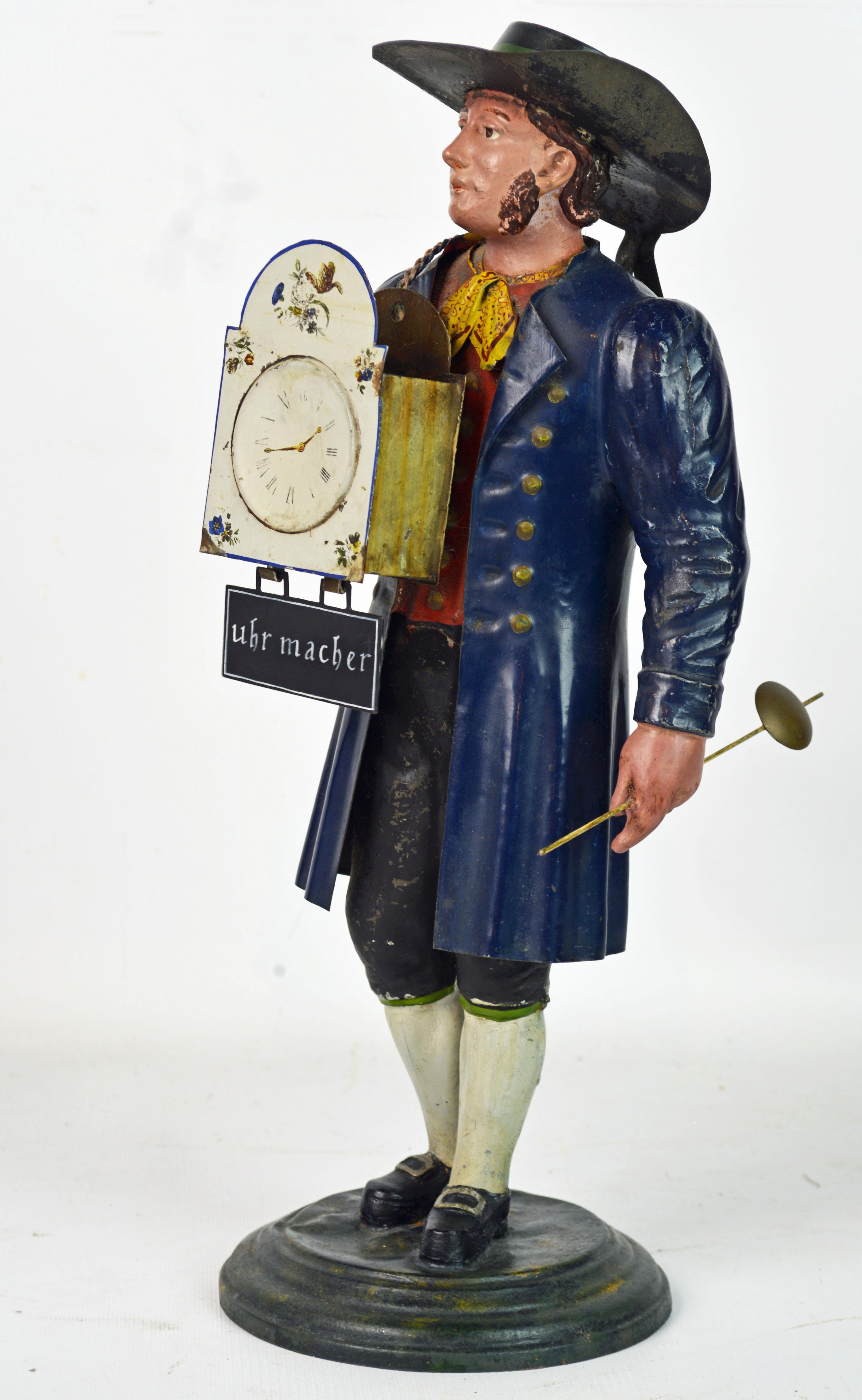 A German Black Forest polychrome painted tole figure of a clock maker in traditional dress, carrying a shield clock in his right and a pendulum in his left hand. Under the clock a sign bearing the inscrition in German 'Uhr Macher'. An interesting