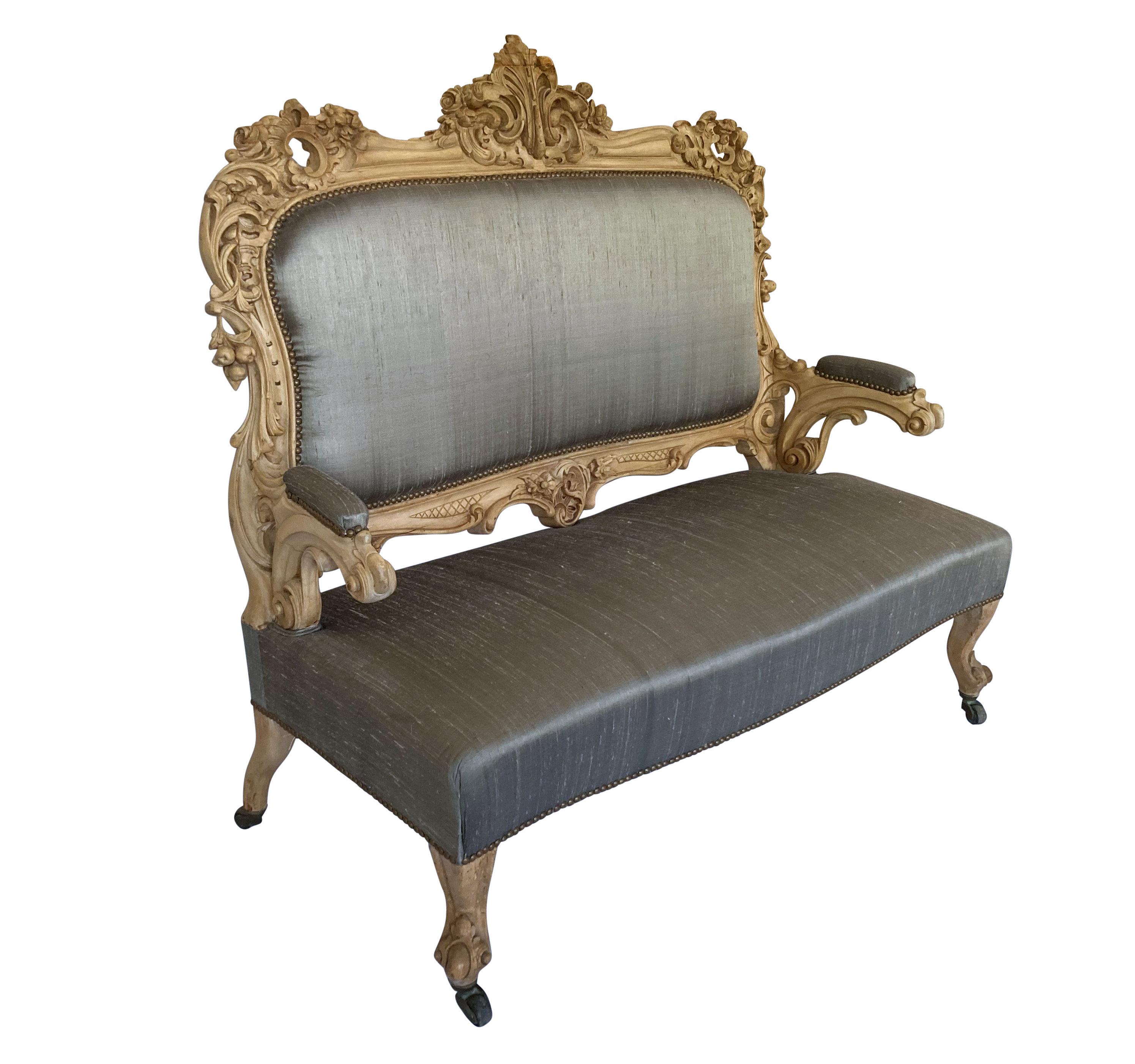 A German early XIX Century settee in the Rococo manner. In beautifully carved bleached fruitwood and upholstered in stone coloured silk with brass studs and brass caster feet.