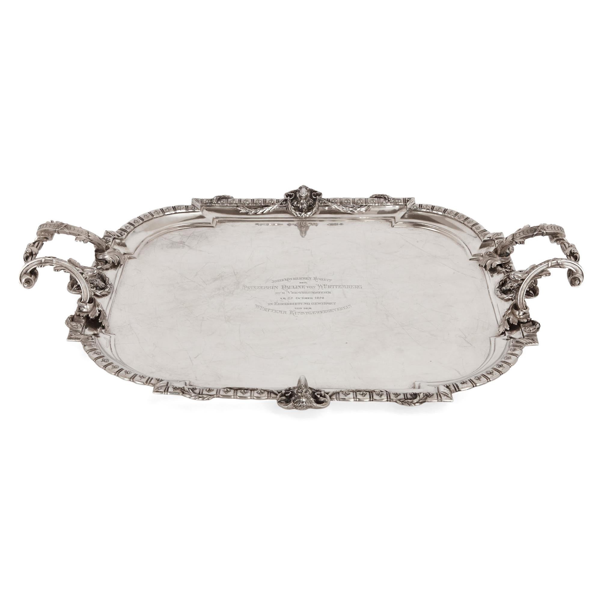 Produced in Germany in the latter decades of the 19th century, this silver tray is rectangular in form with rounded edges and a moulded lip. The tray has two scrolled foliate handles and features a distinctive garlanded ram’s head on each of its