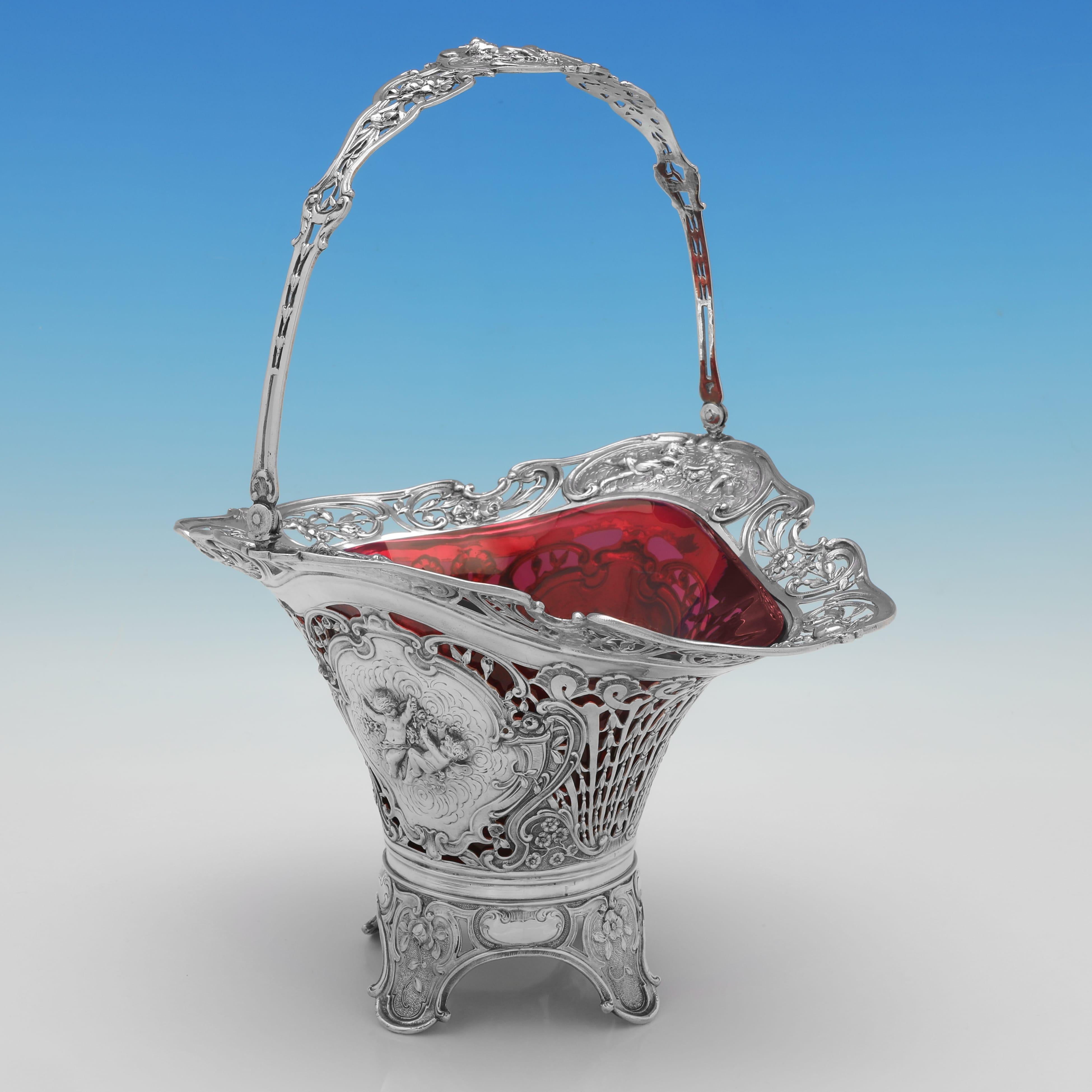 This stunning antique flower basket, crafted in Germany circa 1890, showcases the elegance of 800 standard silver paired with a striking ruby-red glass liner. Featuring ornate pierced work and richly chased repoussé panels, the basket’s design is