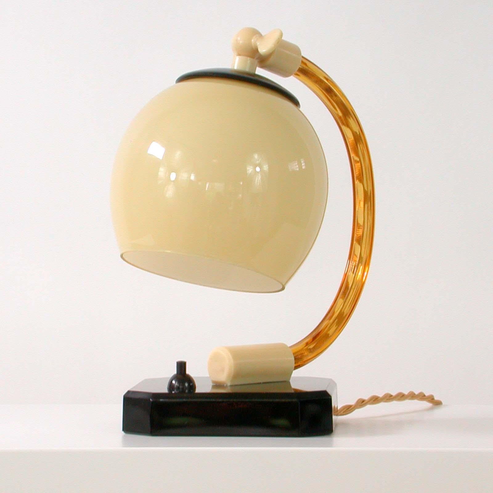 This unusual vintage table or bedside lamp was made in Germany in the 1930s during the Bauhaus period. It is made of black glass (base and top of lamp shade), amber colored clear glass (lamp arm), cream colored bakelite and has got an opaline glass
