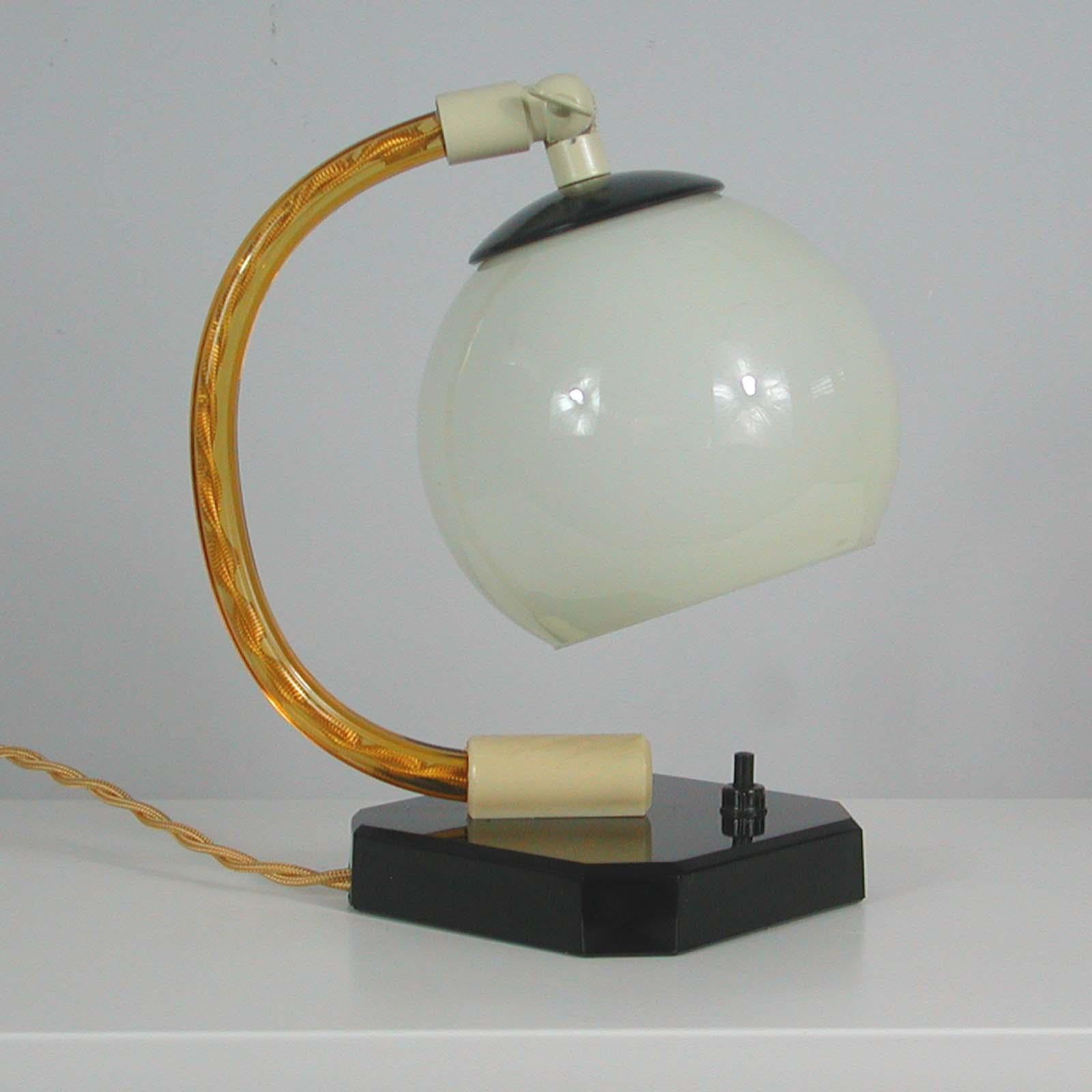 This unusual vintage table or bedside lamp was made in Germany in the 1930s during the Bauhaus period. It features a black glass base and top of lamp shade, amber colored clear glass lamp arm, cream colored bakelite and has got an opaline glass