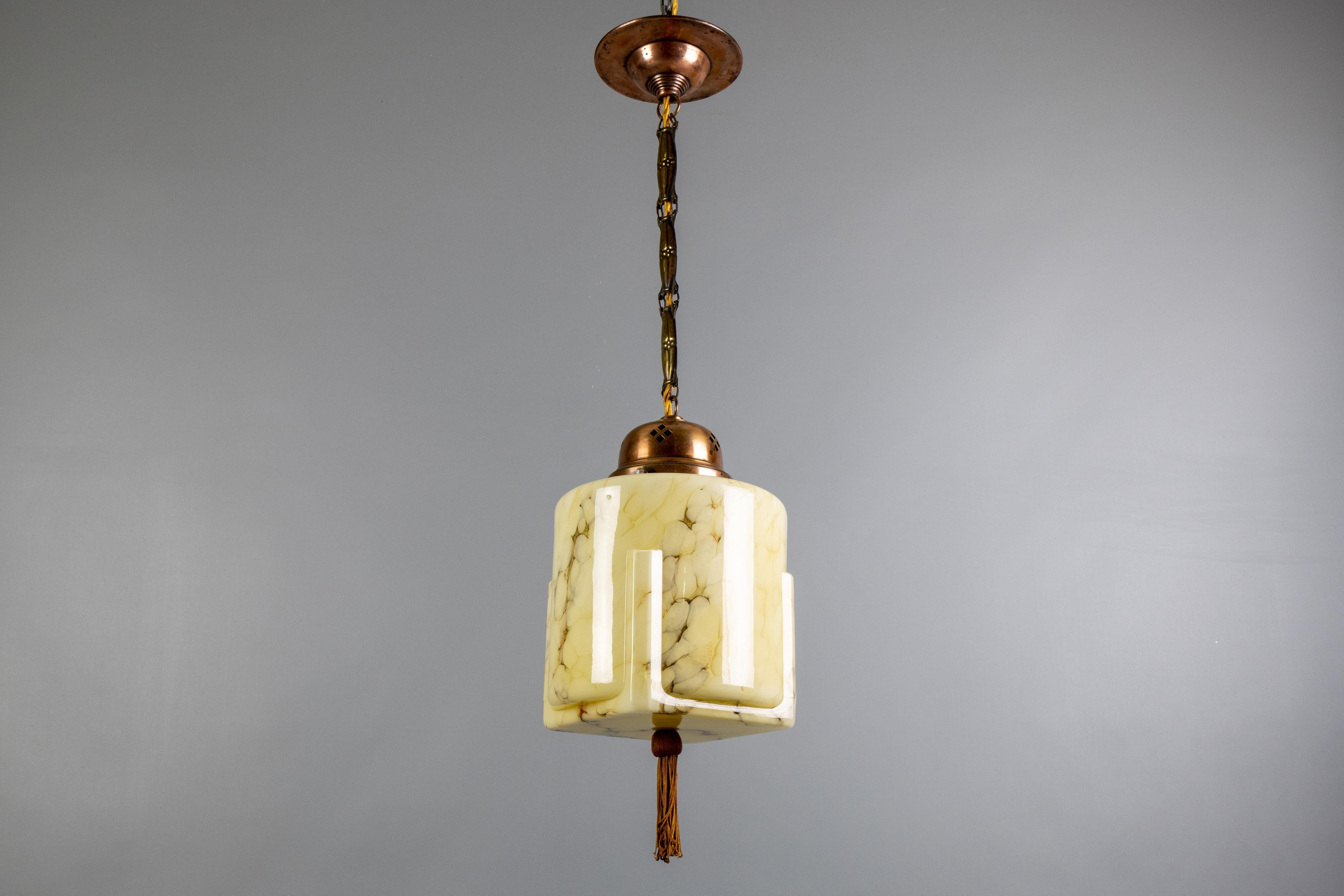 An Art Deco marbled amber color glass and copper pendant light from the 1930s, Germany.
This adorable ceiling light fixture features a beautifully shaped, unusual marbled glass lampshade - round and square simultaneously, hung on a decorative copper